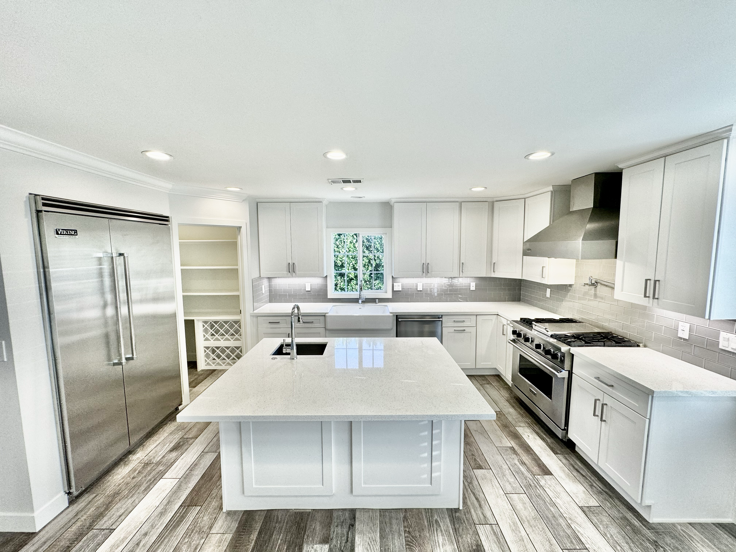 Modern kitchen with white cabinets, stainless steel appliances, a central island with a sink, gray tiled backsplash, and wooden flooring.