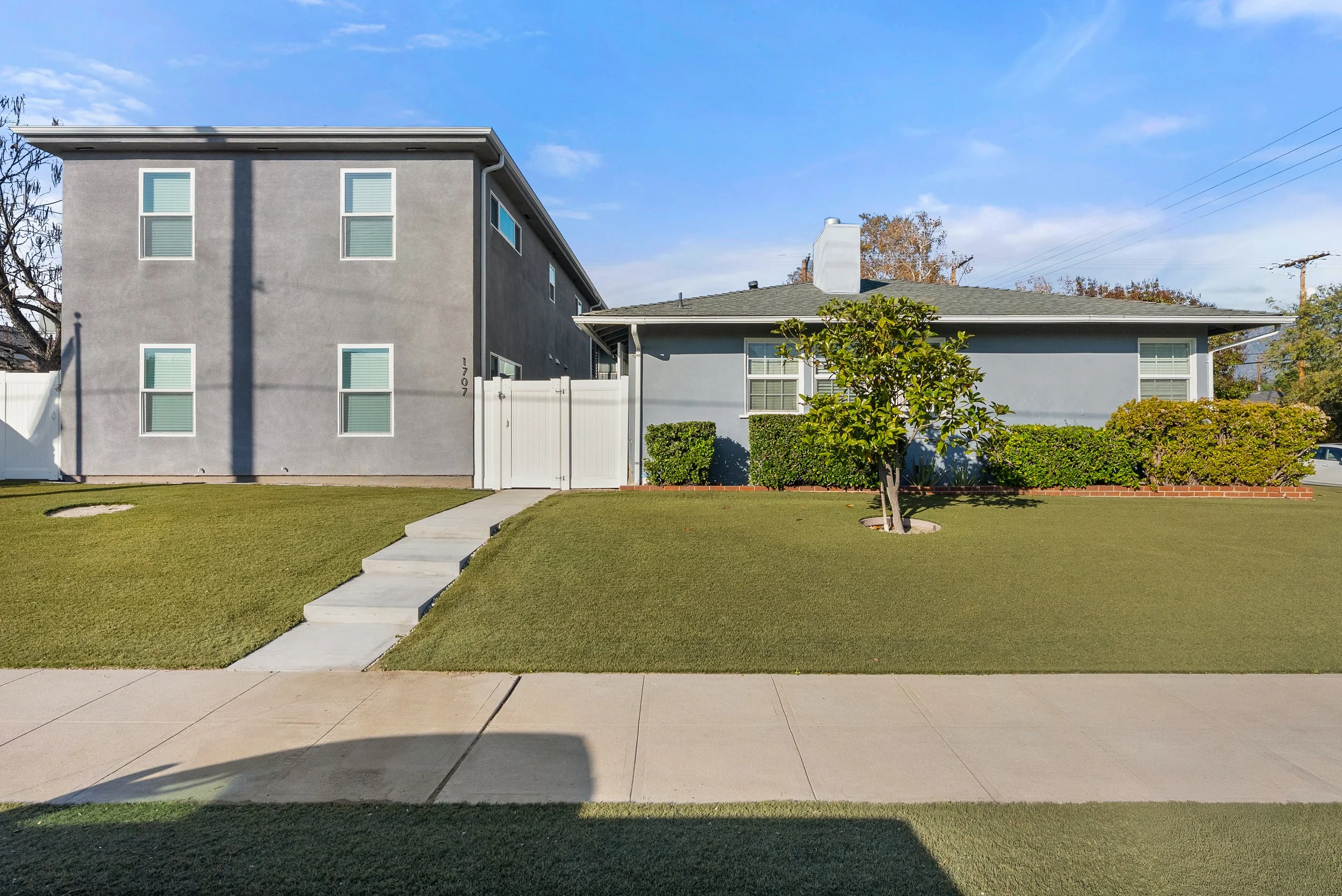 Front view of two houses separated by a white gate, with a sidewalk and lawn in front, and a blue sky overhead.