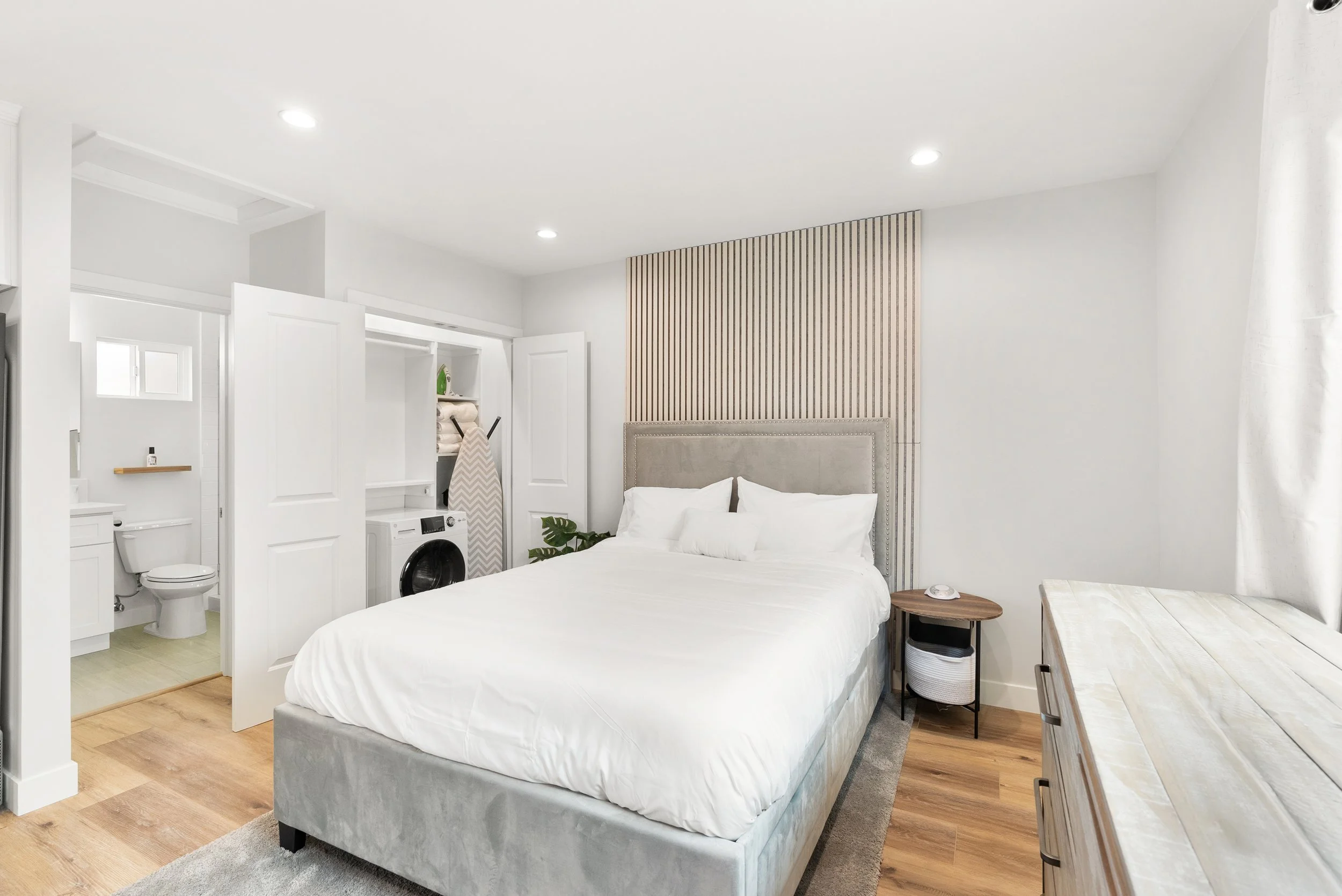 Bedroom with a bed, side table, dresser, laundry area with washing machine, bathroom with toilet, and laundry basket in a closet.