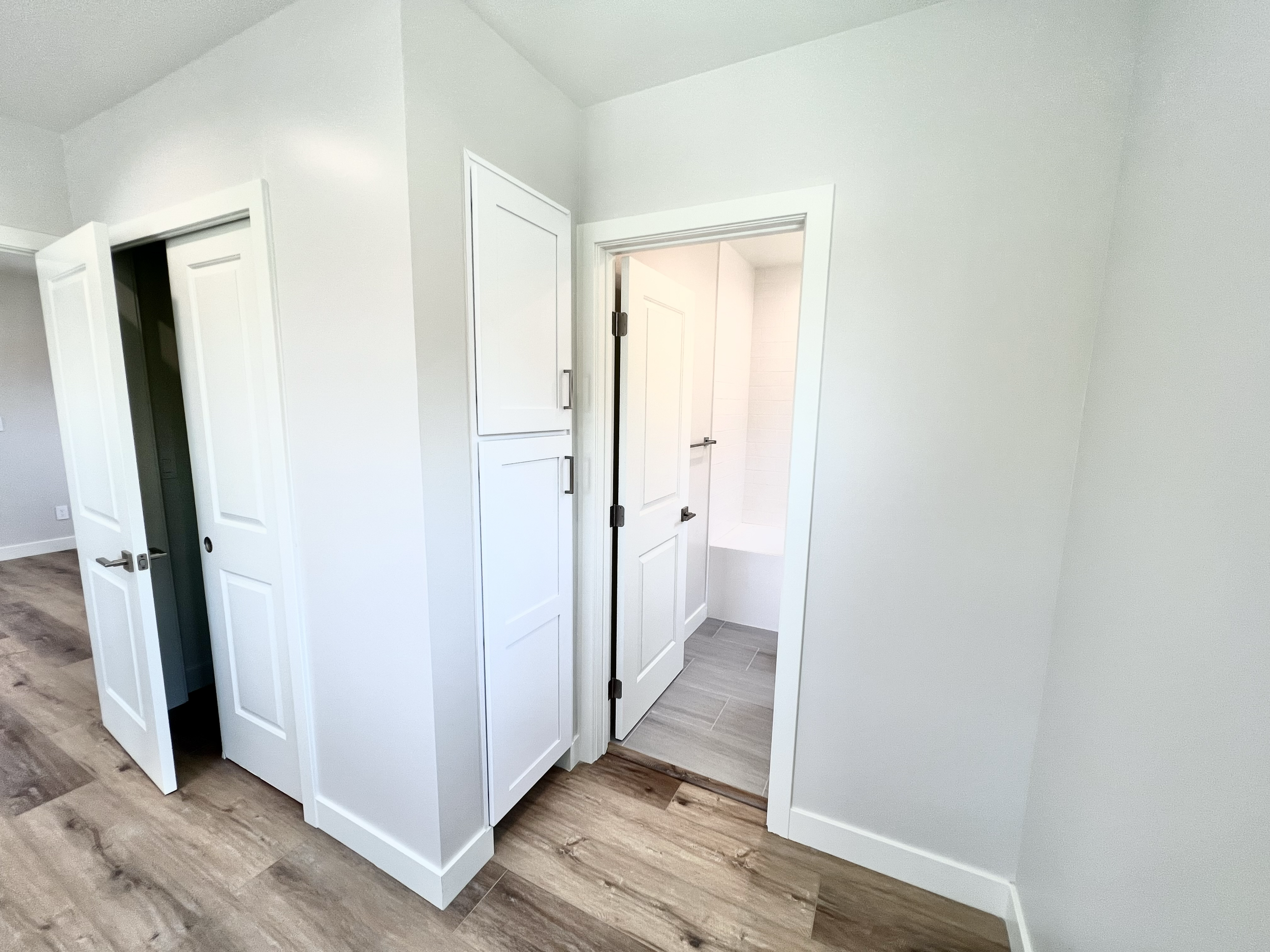Interior view of a modern home bathroom with a partially open door, white cabinetry, and wood-look flooring.