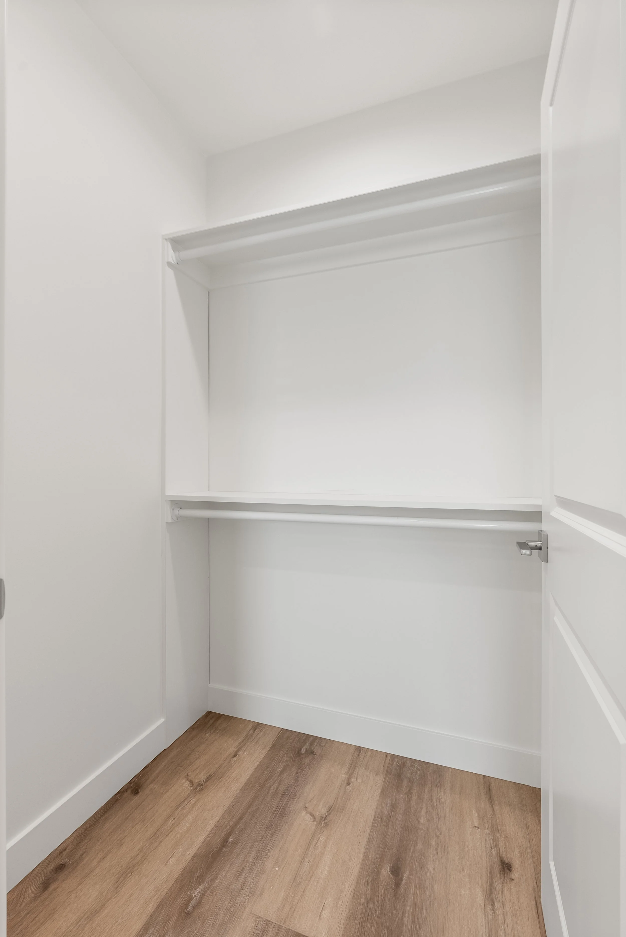Empty white walk-in closet with two shelves and a hanging rod, white walls, wood flooring.