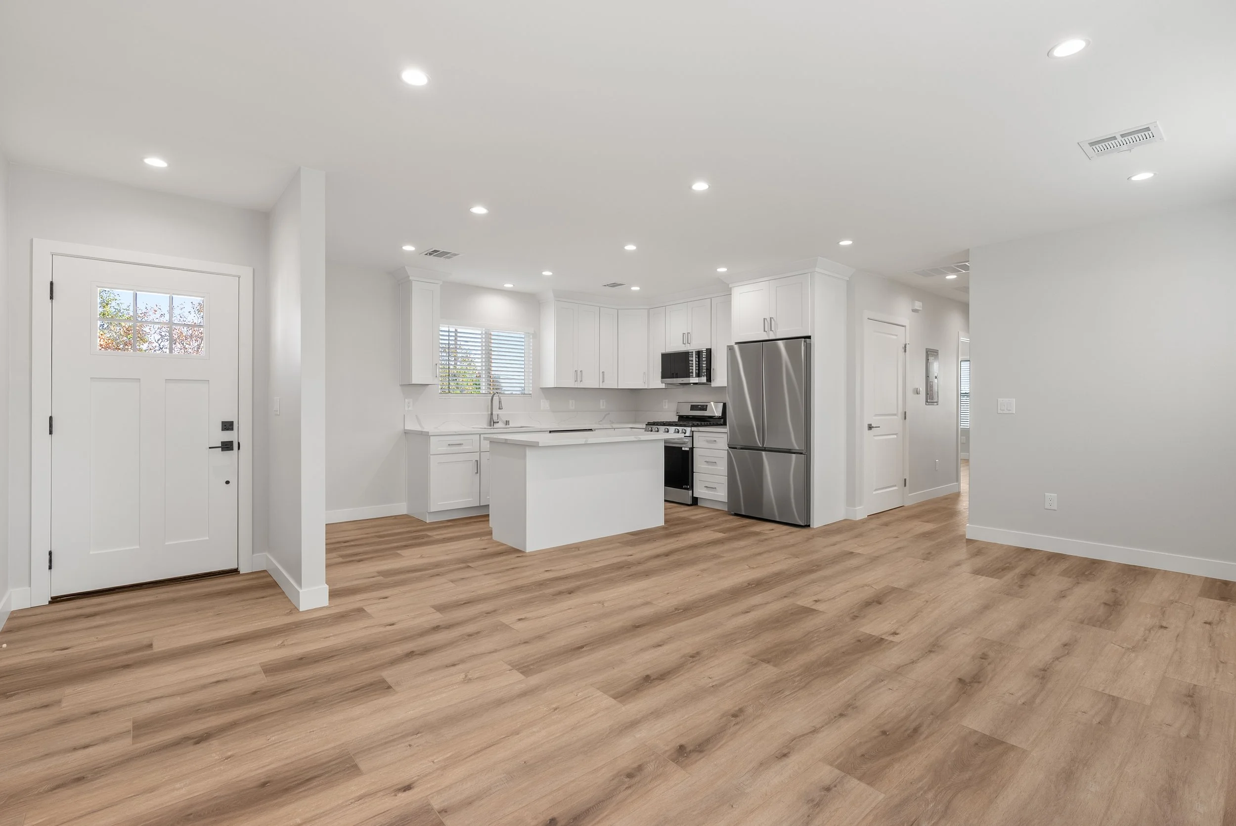 Empty modern kitchen with white cabinets, stainless steel appliances, wood flooring, and a white door.