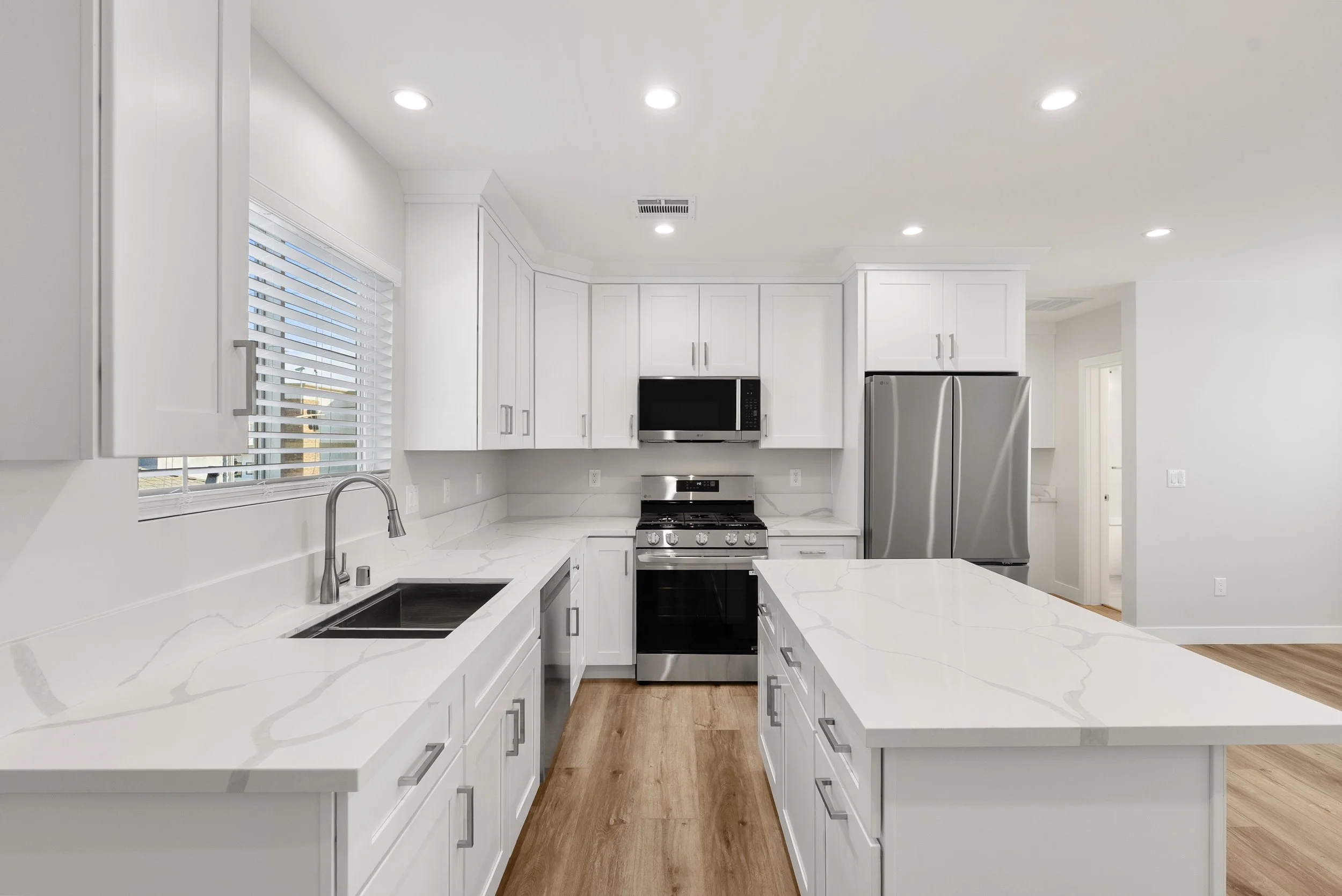 Modern kitchen with white cabinets, marble countertops, stainless steel appliances, a window with blinds, wood flooring, and recessed lighting.
