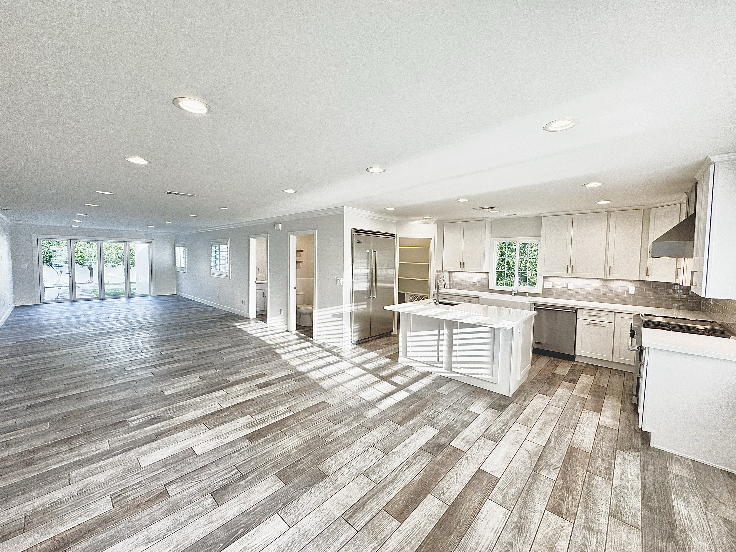 Open-plan living area and kitchen with wood-look flooring, white cabinetry, stainless steel appliances, and large windows letting in natural light.