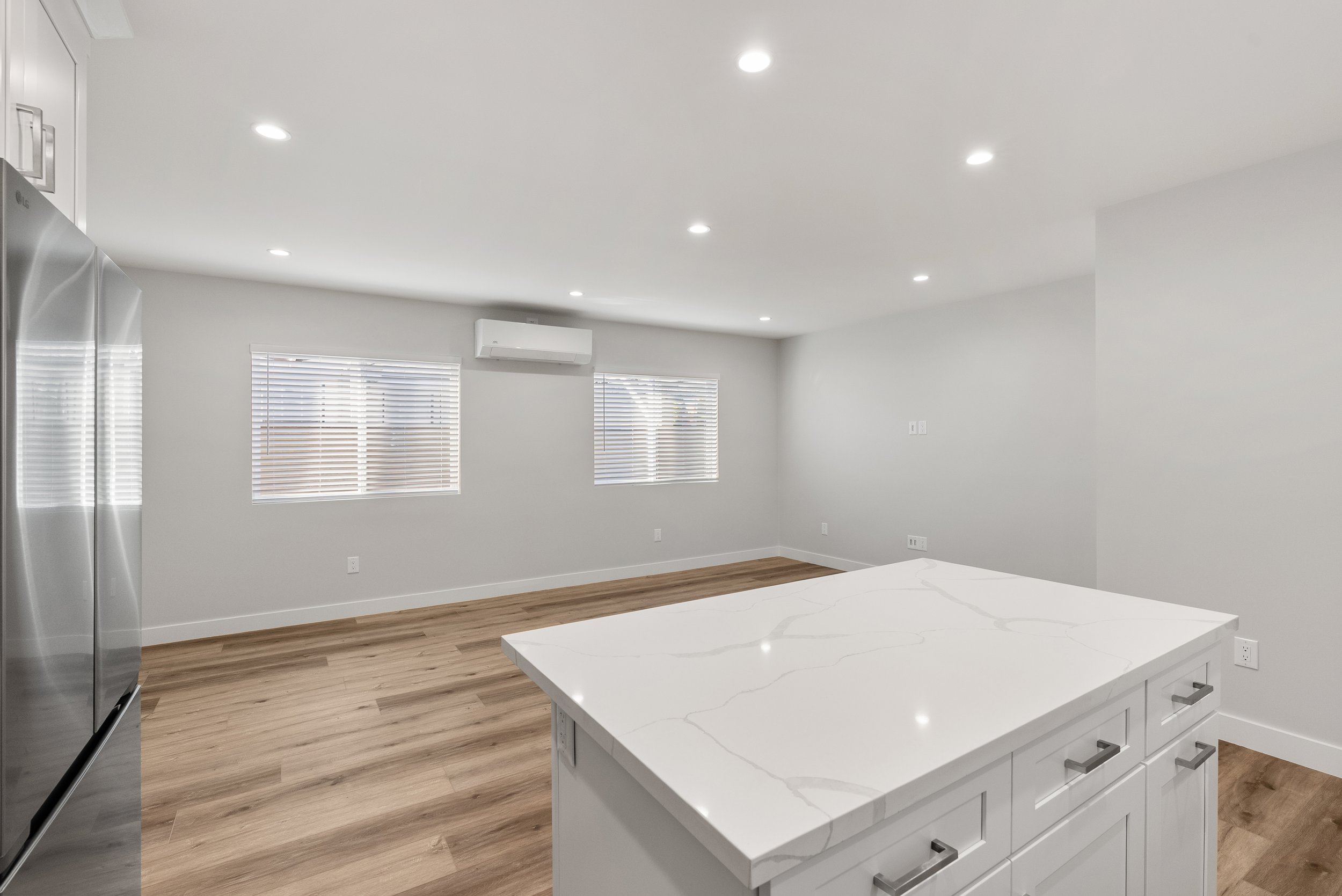 Empty modern kitchen with white marble island, stainless steel refrigerator, hardwood floors, and white walls with windows and an air conditioning unit.