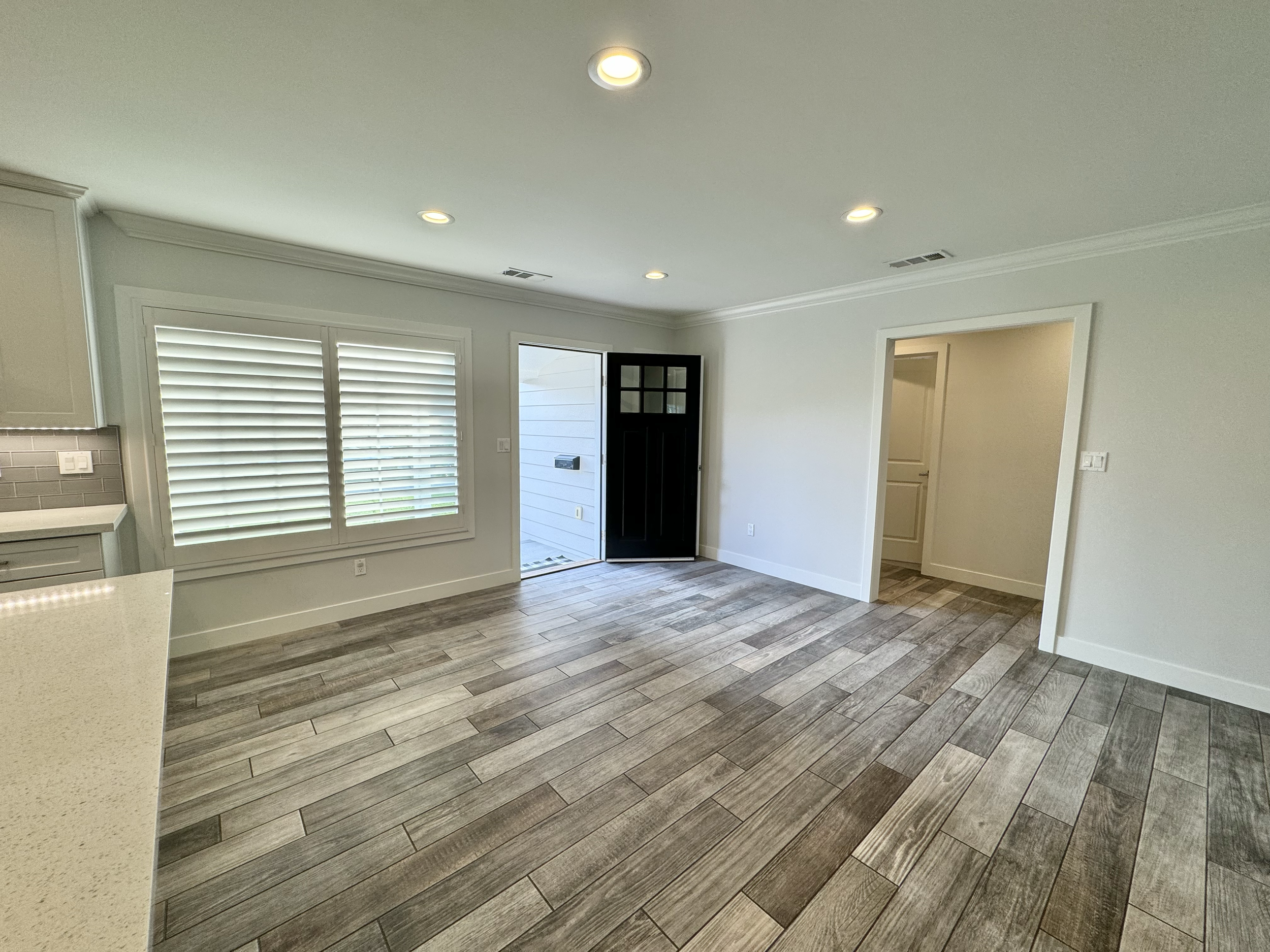 Empty living room with wood flooring, white walls, white window shutters, and a black front door, with a small hallway visible through an open doorway.