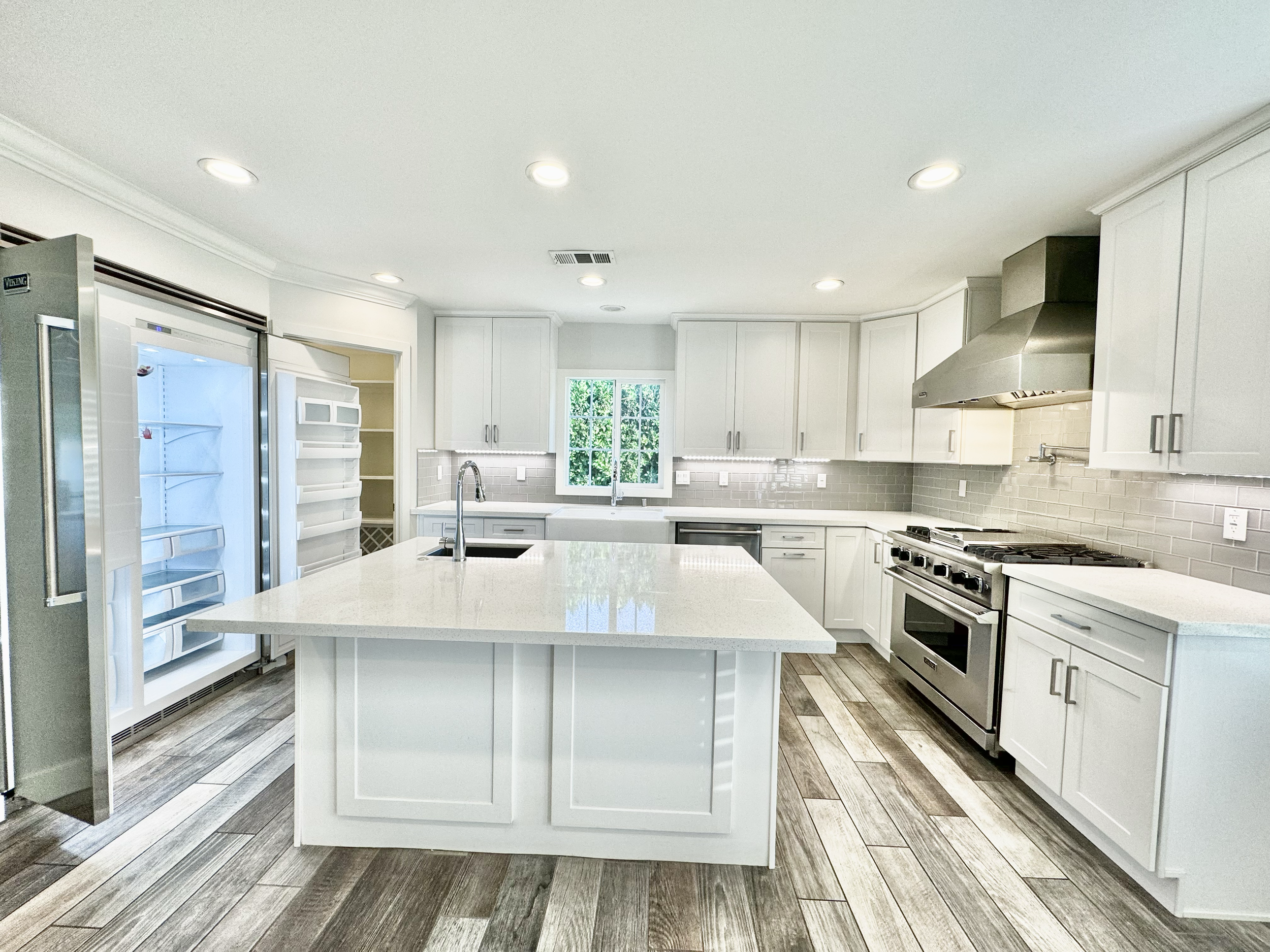 Modern kitchen with white cabinets, stainless steel appliances, island with sink, gray backsplash, wooden flooring, and a window over the sink.