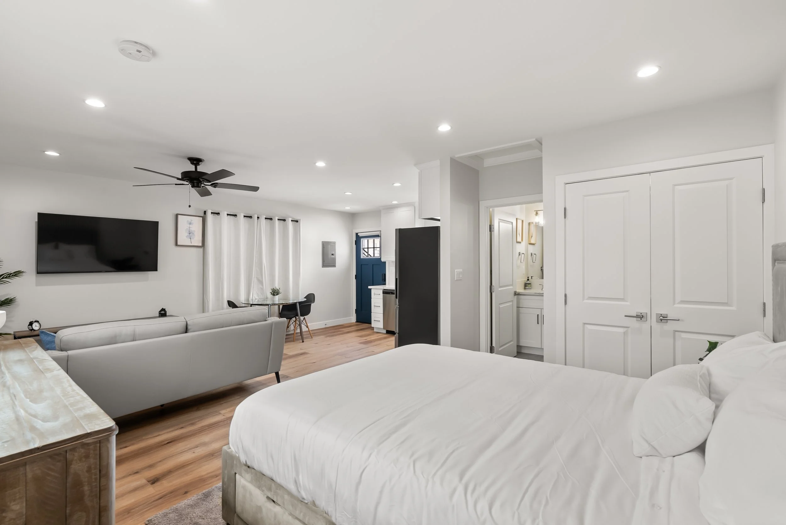 A spacious, modern bedroom with white walls and ceiling, a large bed with white linens, a flat-screen TV on the wall, a ceiling fan, hardwood floors, and a small sitting area with a table and chairs near a window with white curtains.