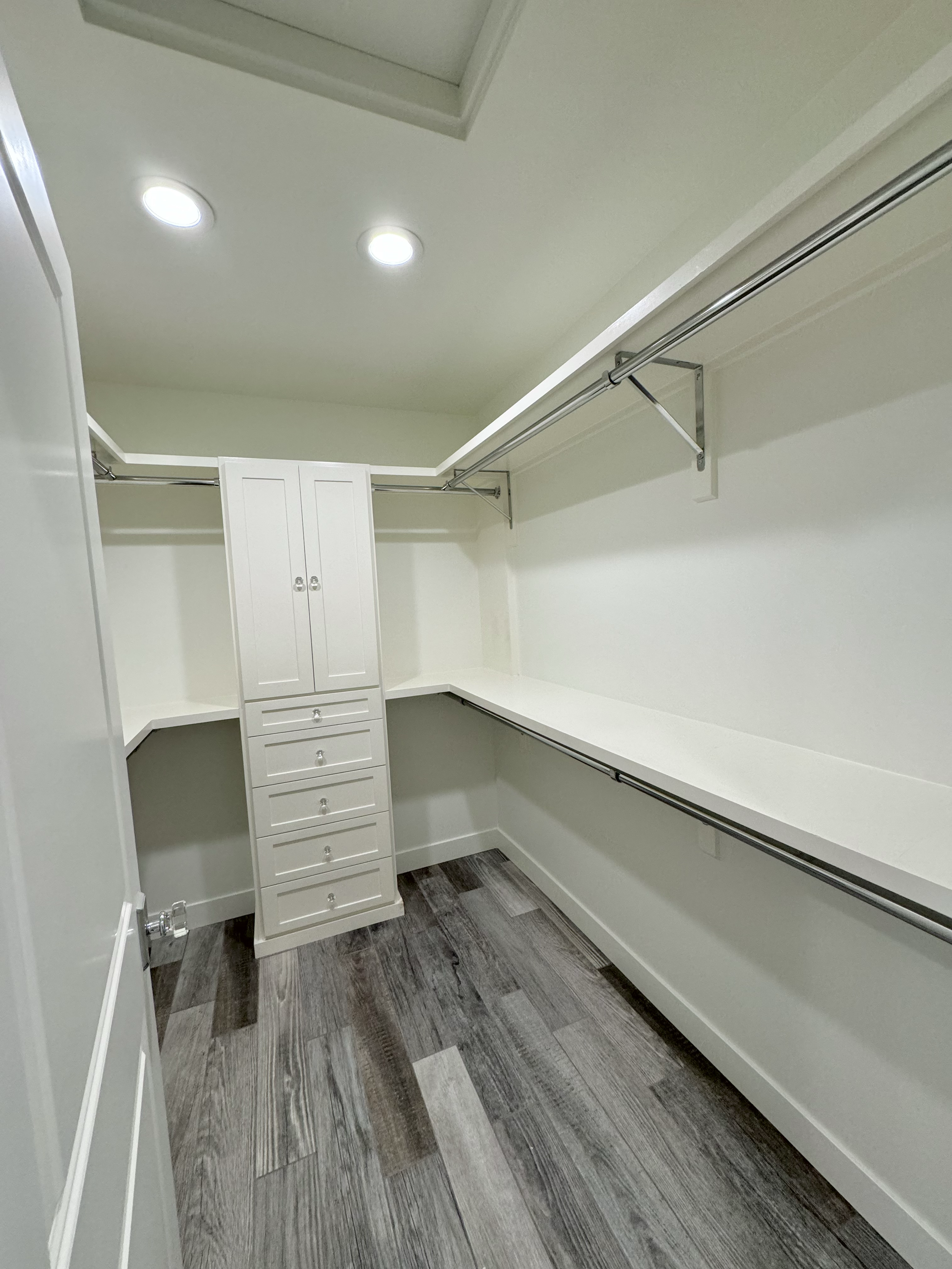Empty walk-in closet with white shelves, drawers, and hanging rods, and dark wood flooring.