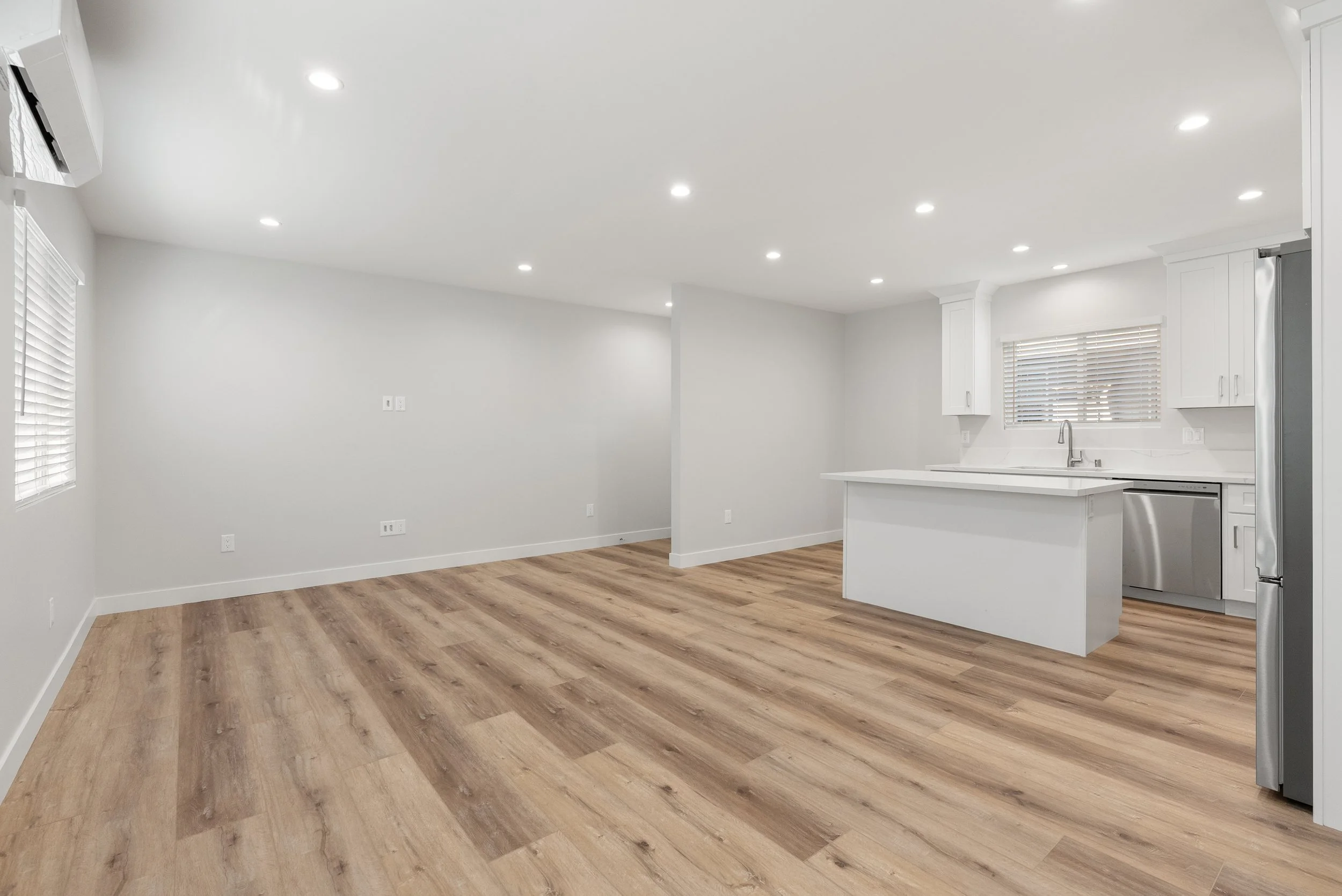 Empty modern kitchen and living area with white walls, hardwood floors, white cabinetry, stainless steel appliances, and recessed lighting.