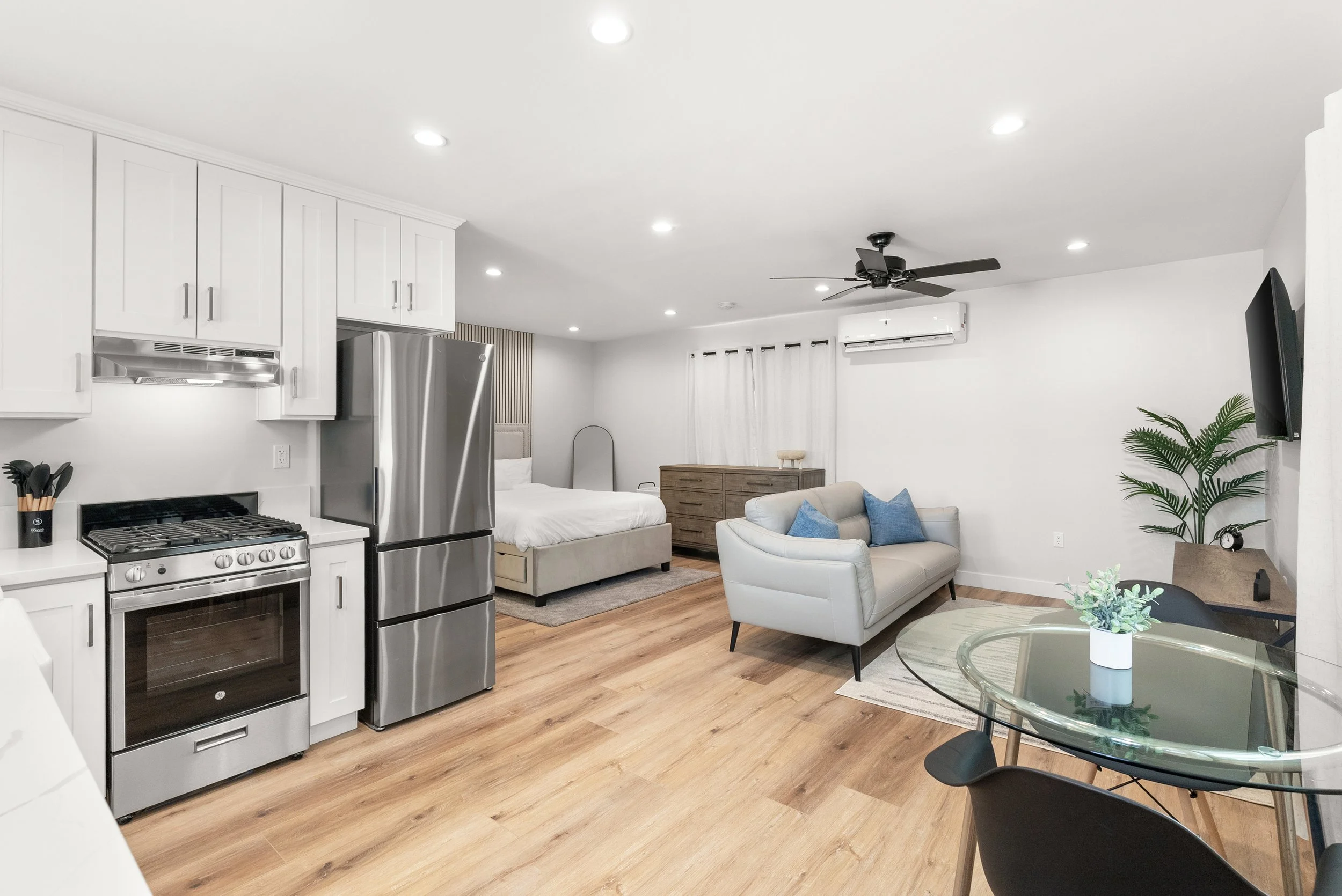 Open-plan living space with kitchen, dining, and sleeping area. The kitchen has white cabinets, a stainless steel refrigerator, stove, and oven. The room has wood flooring, a white sofa with blue pillows, a bed with white bedding, a wooden dresser, a