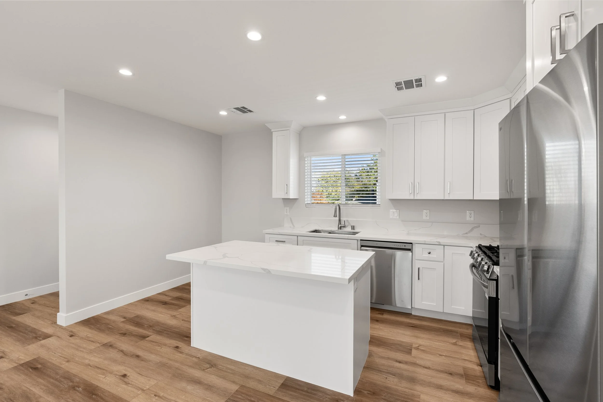 Modern kitchen with white cabinets, marble countertops, stainless steel appliances, wooden flooring, window with blinds, and ceiling lights.