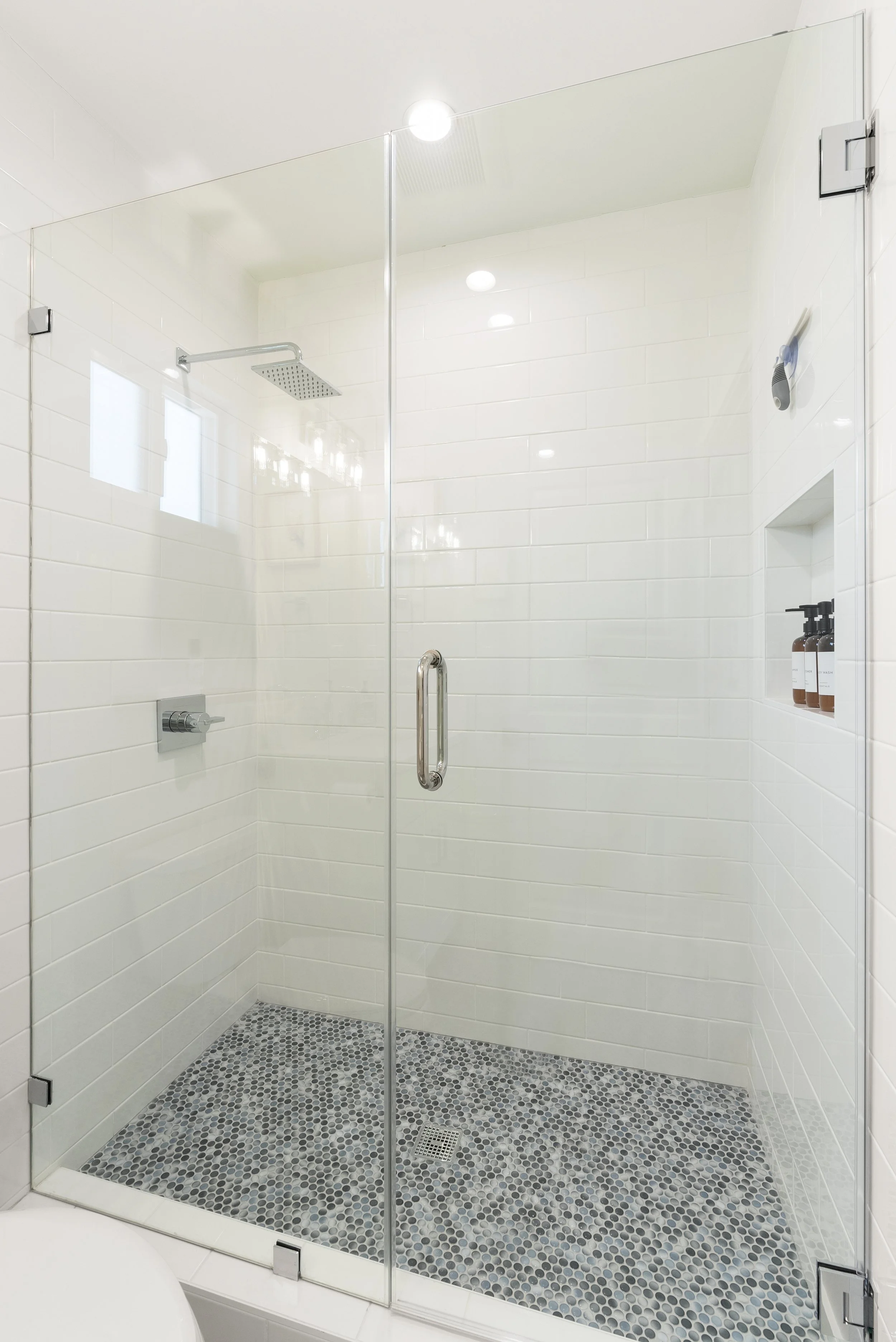 Minimalist glass-enclosed shower with white subway tile walls, pebble tile floor, built-in shampoo niche, and showerhead