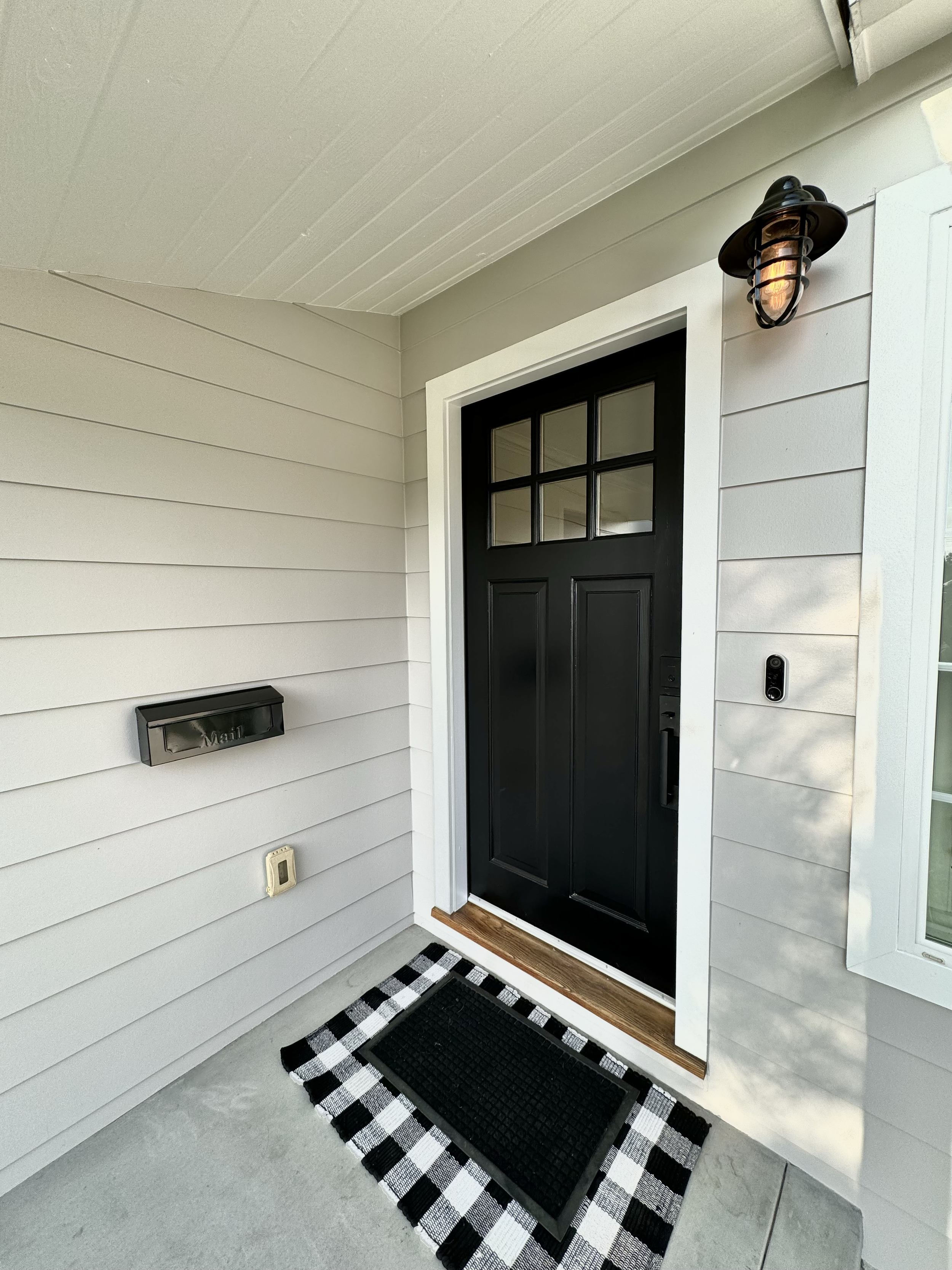 Close-up image of a house front door with a black handle and a small window panel at the top, reached by a doormat with a black and white checkered border and black inner mat, attached to a concrete porch with a wall-mounted mailbox and a porch-style