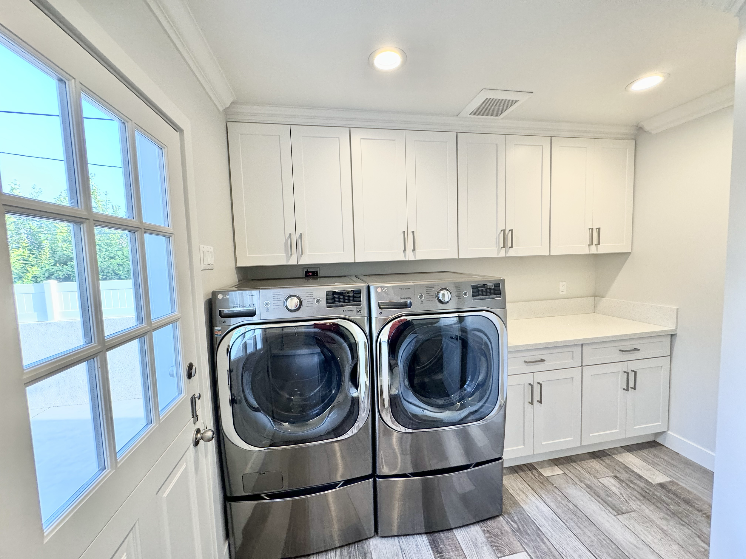 Laundry room with white cabinets, a countertop, and stainless steel LG washer and dryer side by side, with a window showing an outdoor fence and trees.