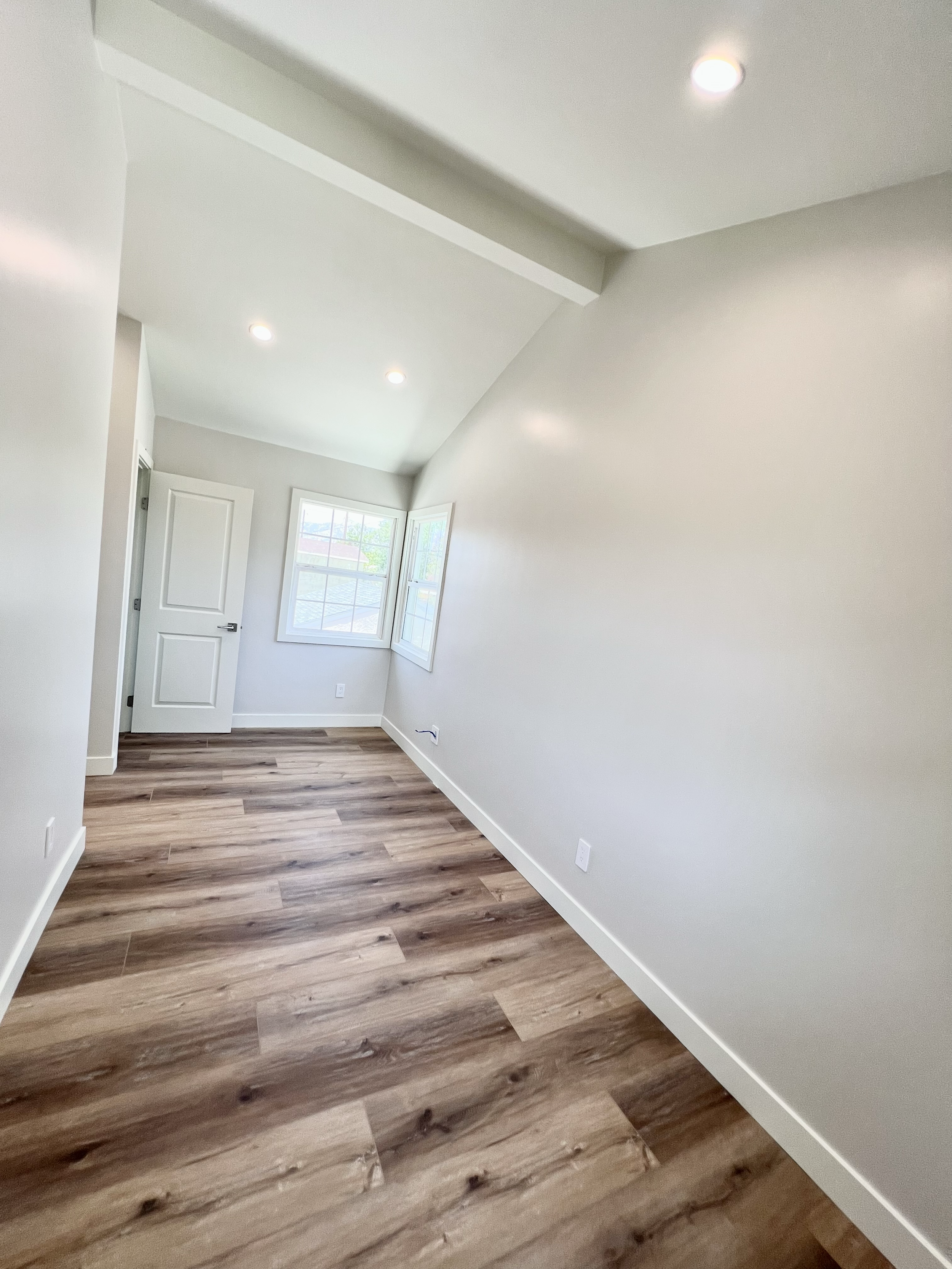 Empty room with wood flooring, white walls, a window, and a door, under a sloped ceiling with recessed lighting.