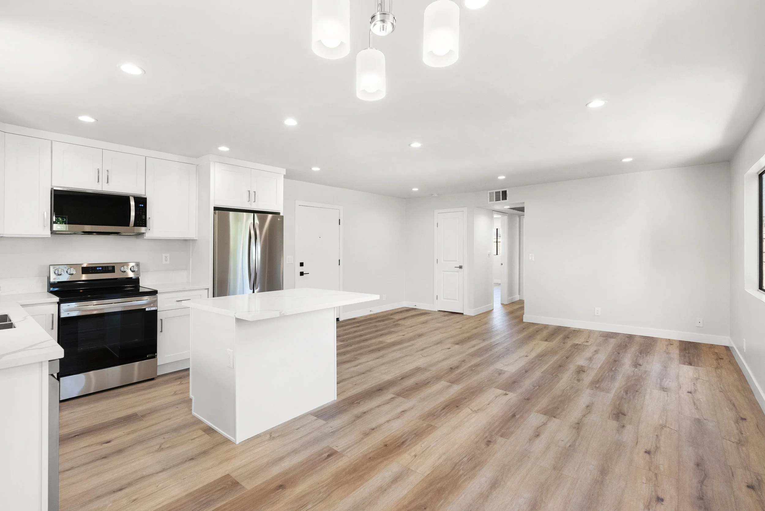 Empty modern kitchen with white cabinets, stainless steel appliances, a white island, hardwood floors, and white walls, with ceiling lights.