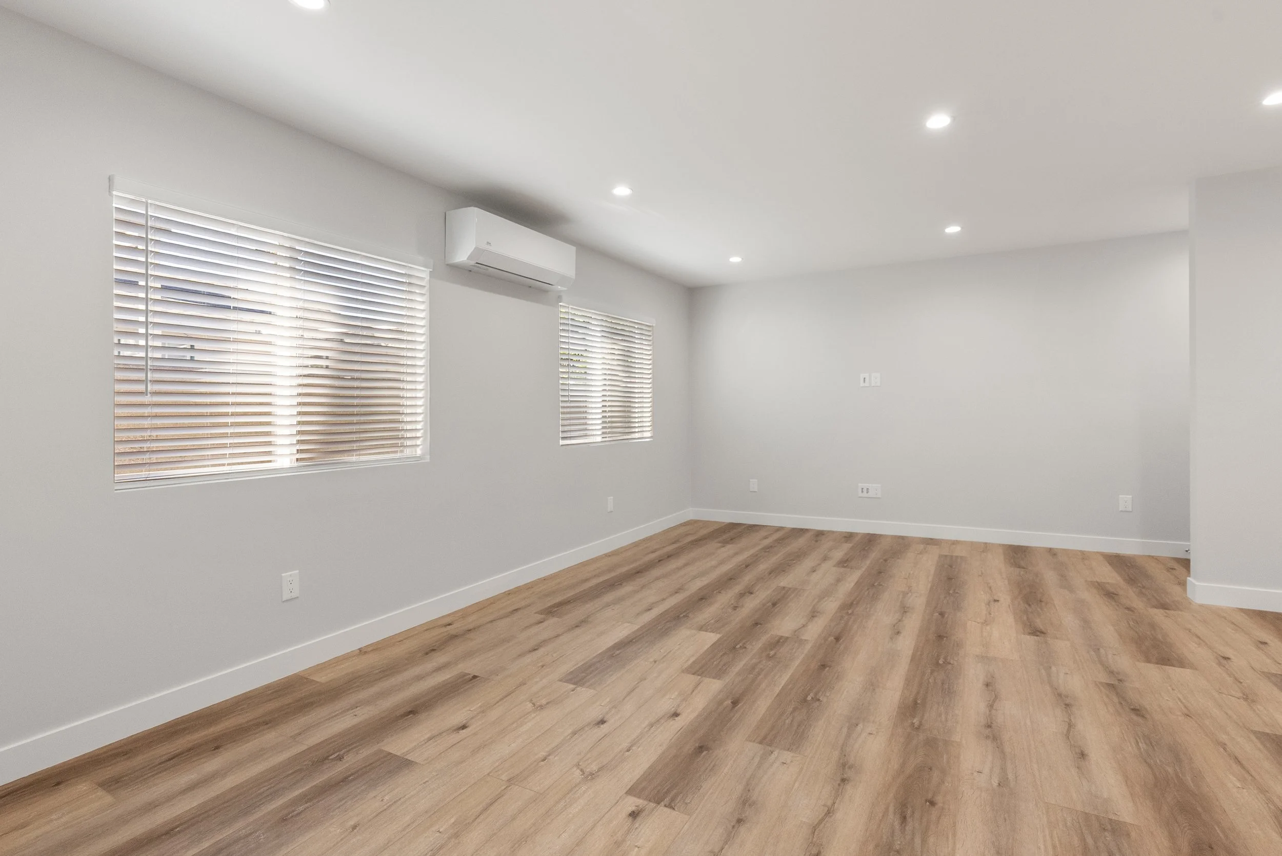 Empty living room with white walls, wooden flooring, two windows with white blinds, and ceiling lights.