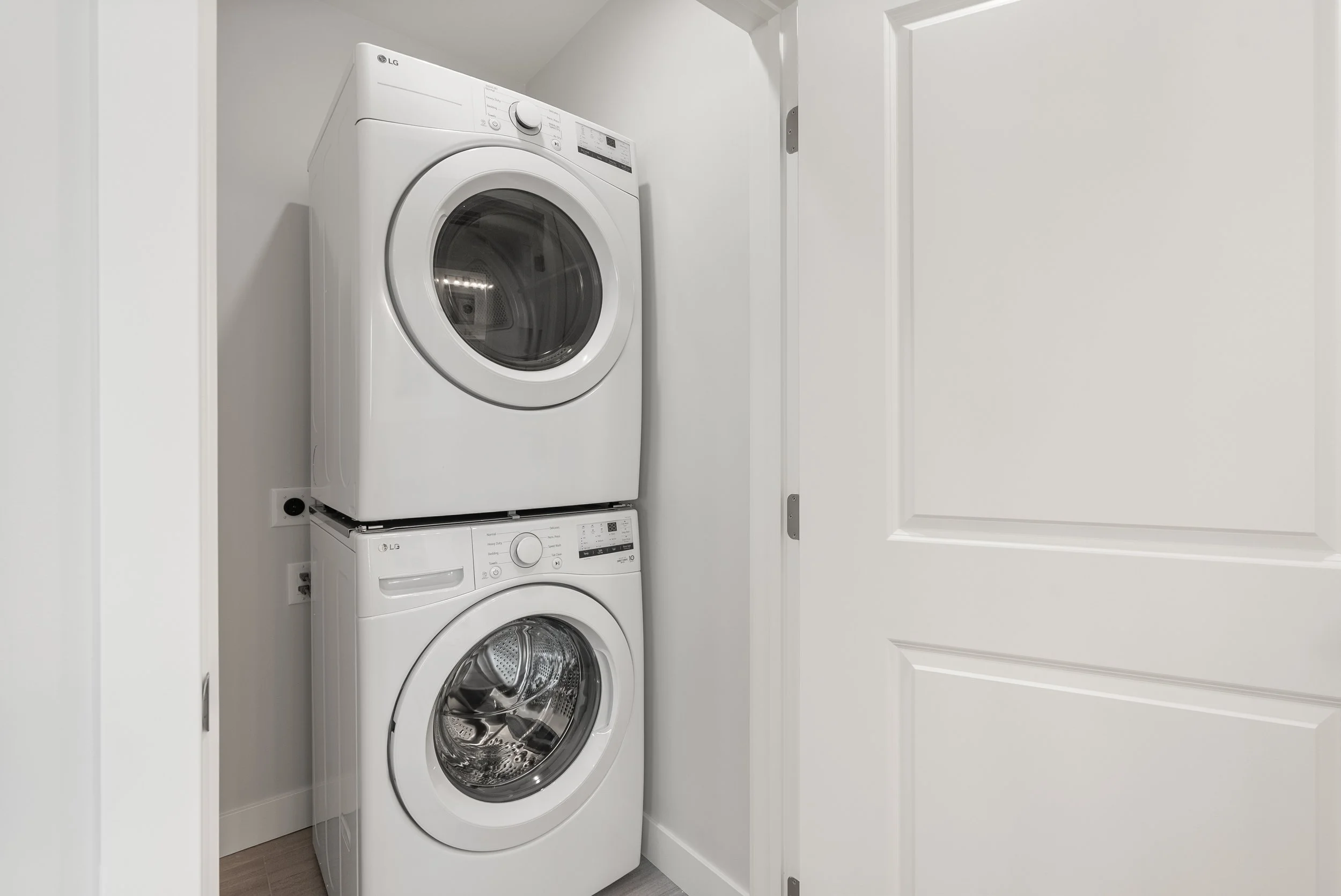 Stacked white front-loading washing machine and dryer in a laundry closet with white walls and door.