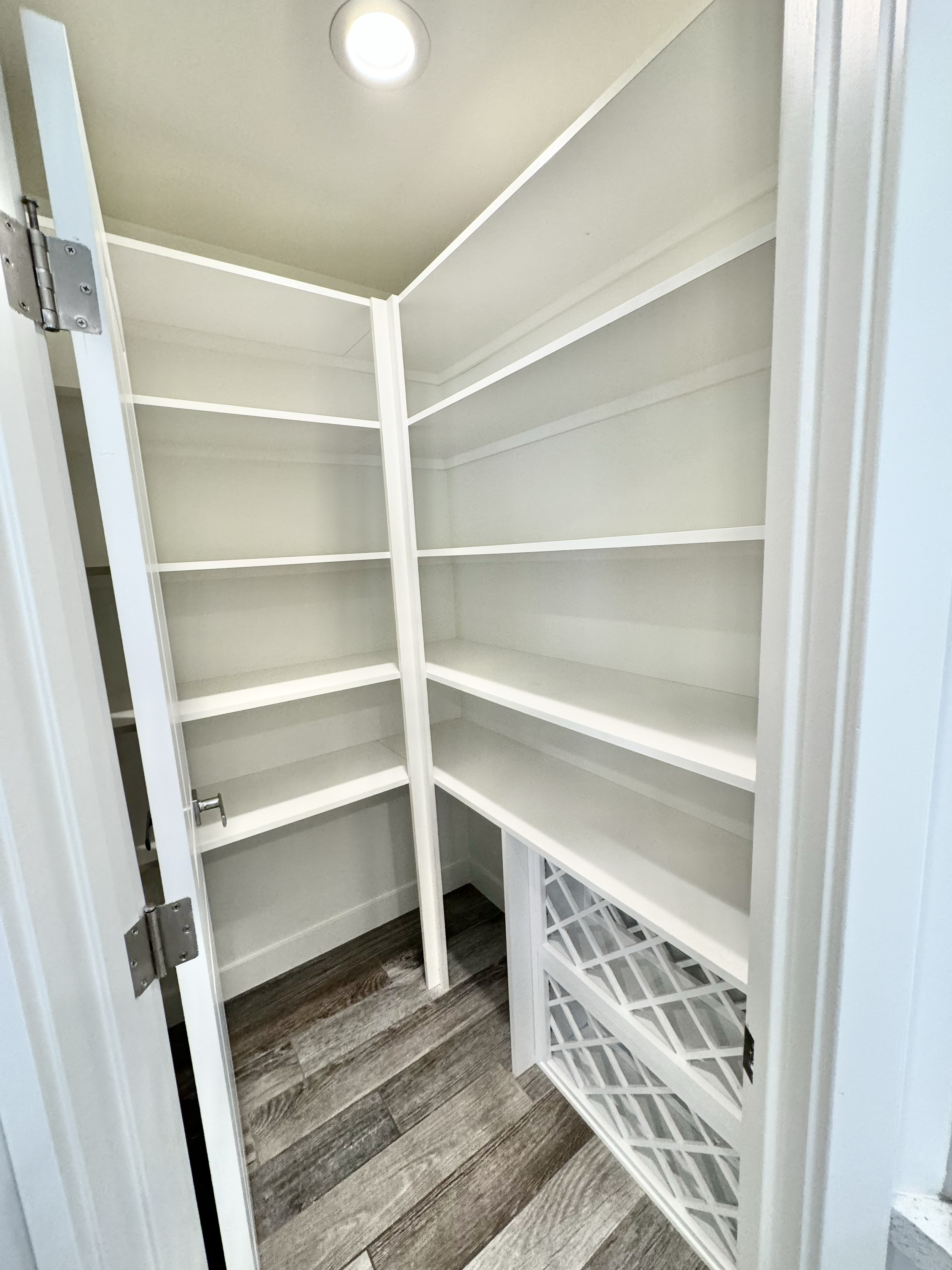 Empty walk-in closet with white shelves and a wine rack at the bottom, brown wooden flooring, and a ceiling light.