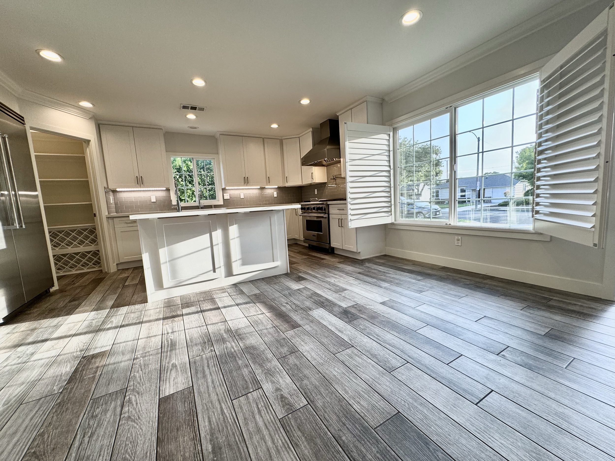 Bright, modern kitchen with wood-look tile flooring, white cabinets, a large window with white shutters, stainless steel appliances, and a kitchen island.