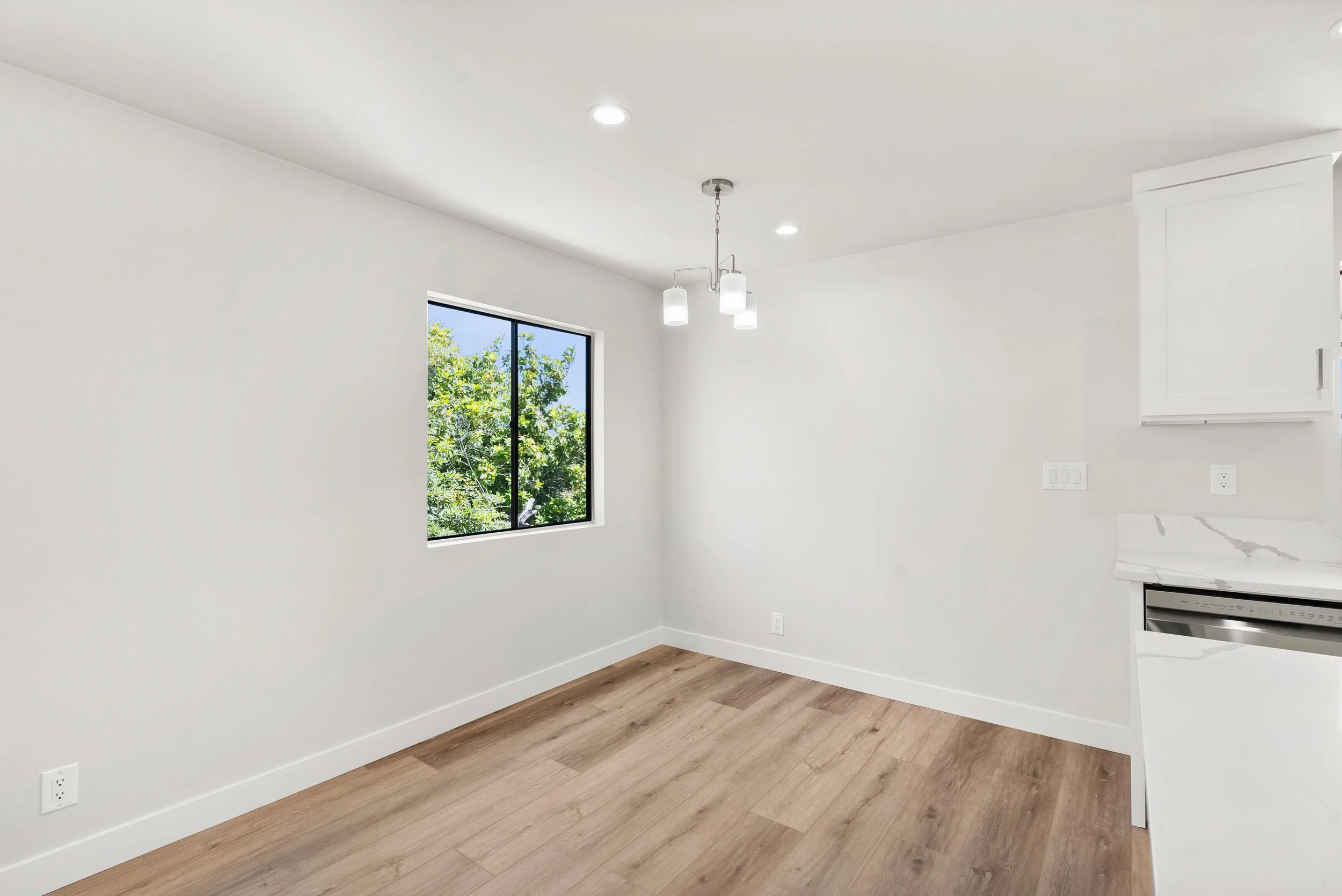 Empty kitchen corner with a window showing green trees outside, white walls, wooden floor, and a hanging ceiling light fixture.