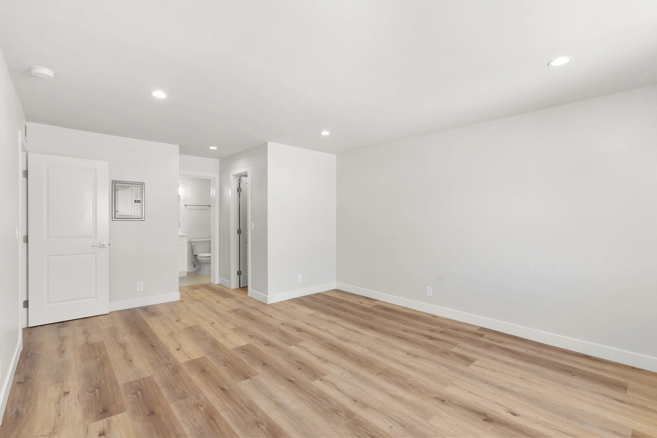 Empty room with white walls, wood flooring, and ceiling lights, with a door leading to a bathroom.