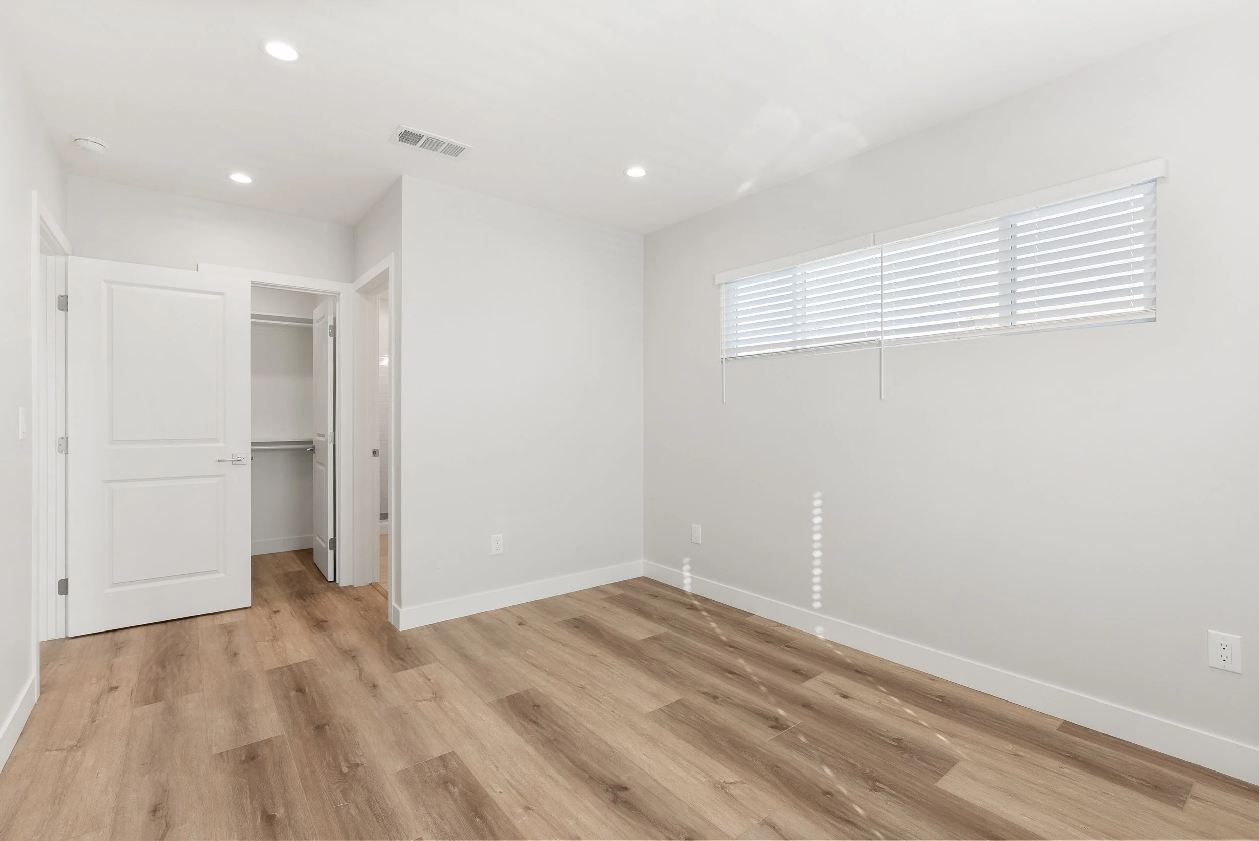 Empty room with hardwood floors, white walls, a window with blinds, and an open closet.
