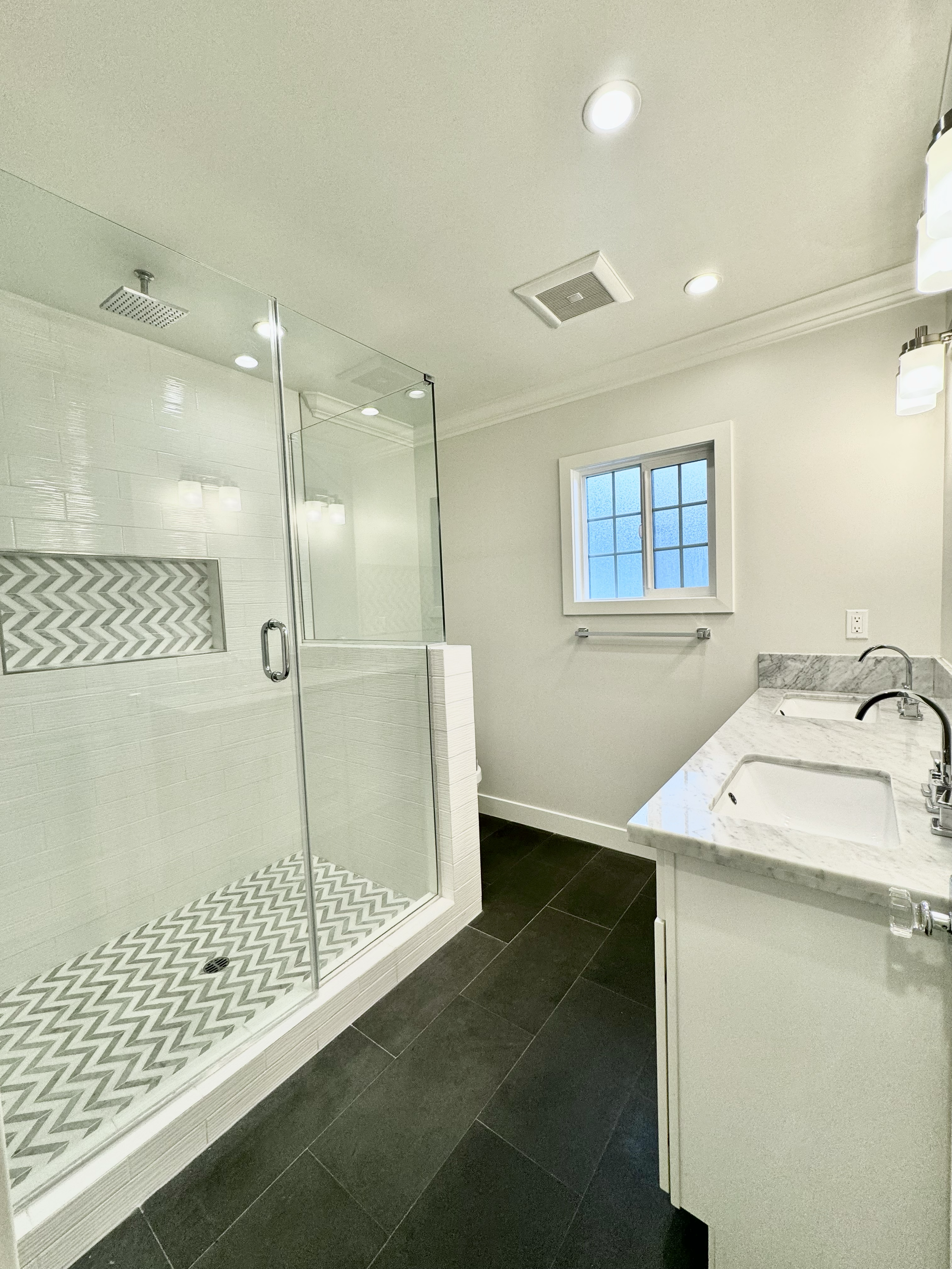 A modern bathroom with a glass-enclosed shower, white tiled walls, a window, and a marble double sink vanity.