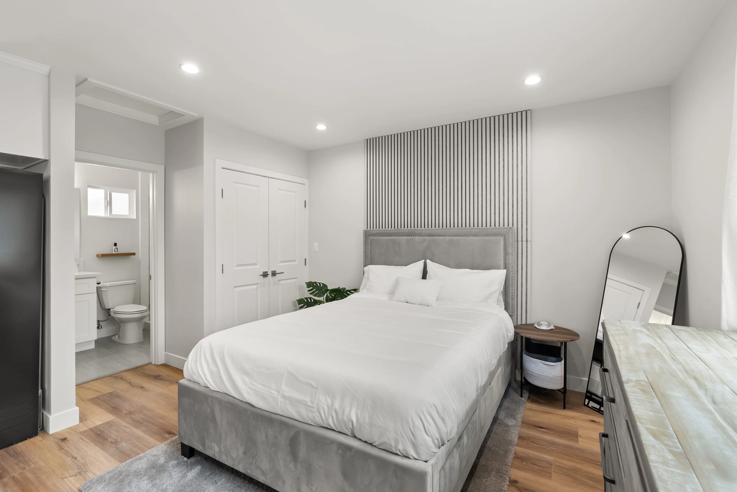 A modern bedroom with a king-sized bed, gray velvet headboard, white bedding, small wooden side table, full-length mirror, and a view into a bathroom with a toilet and small window.
