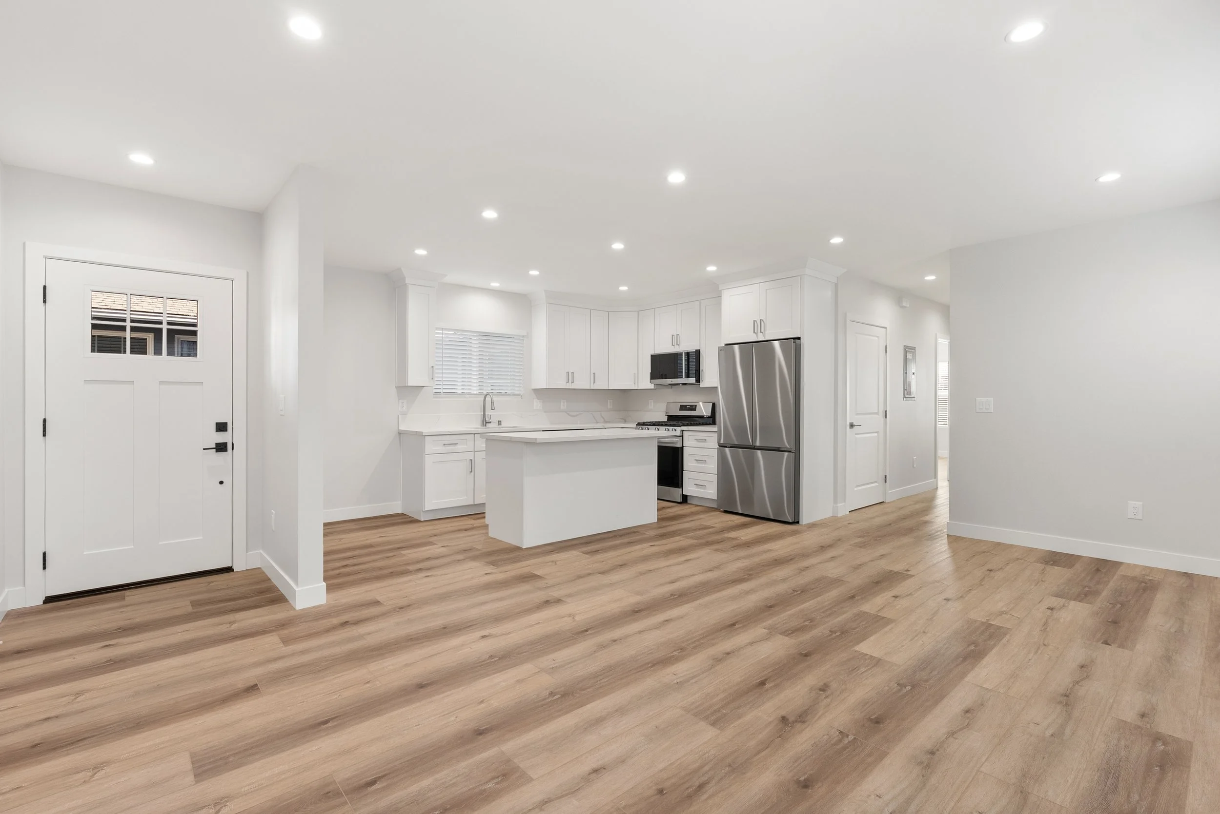 Modern white kitchen with stainless steel refrigerator, microwave, and oven, white cabinets, a window with blinds, a central island, and hardwood flooring.