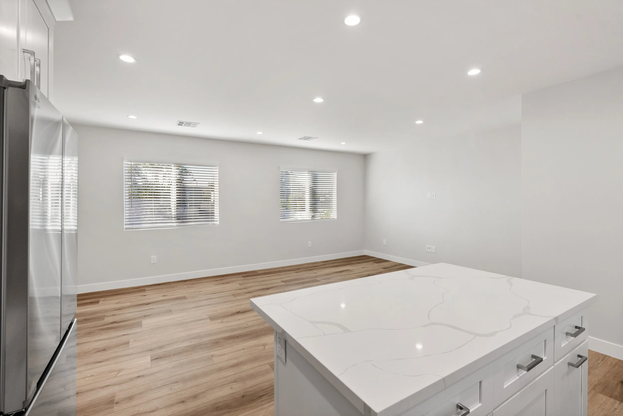 Empty modern kitchen with white walls, wooden floor, stainless steel refrigerator, white countertop island, and two windows with blinds.