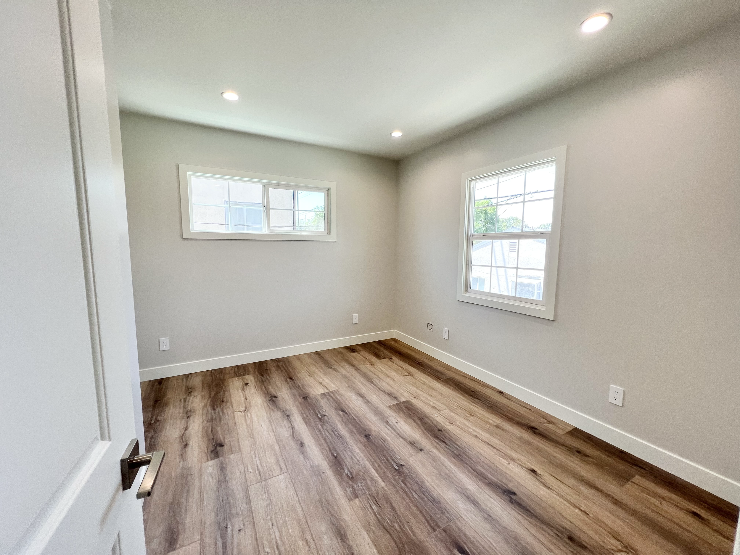 Empty room with white walls, wood flooring, two windows with white frames, and recessed ceiling lights.