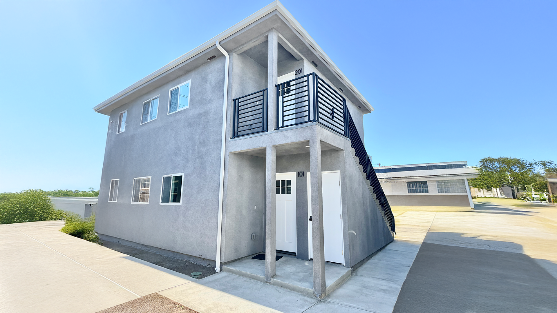 A modern two-story apartment building with gray exterior walls, multiple windows, and a staircase on the right side leading to the upper level. The building has front doors numbered 101 and 201, and is surrounded by a paved area and some greenery in the background.