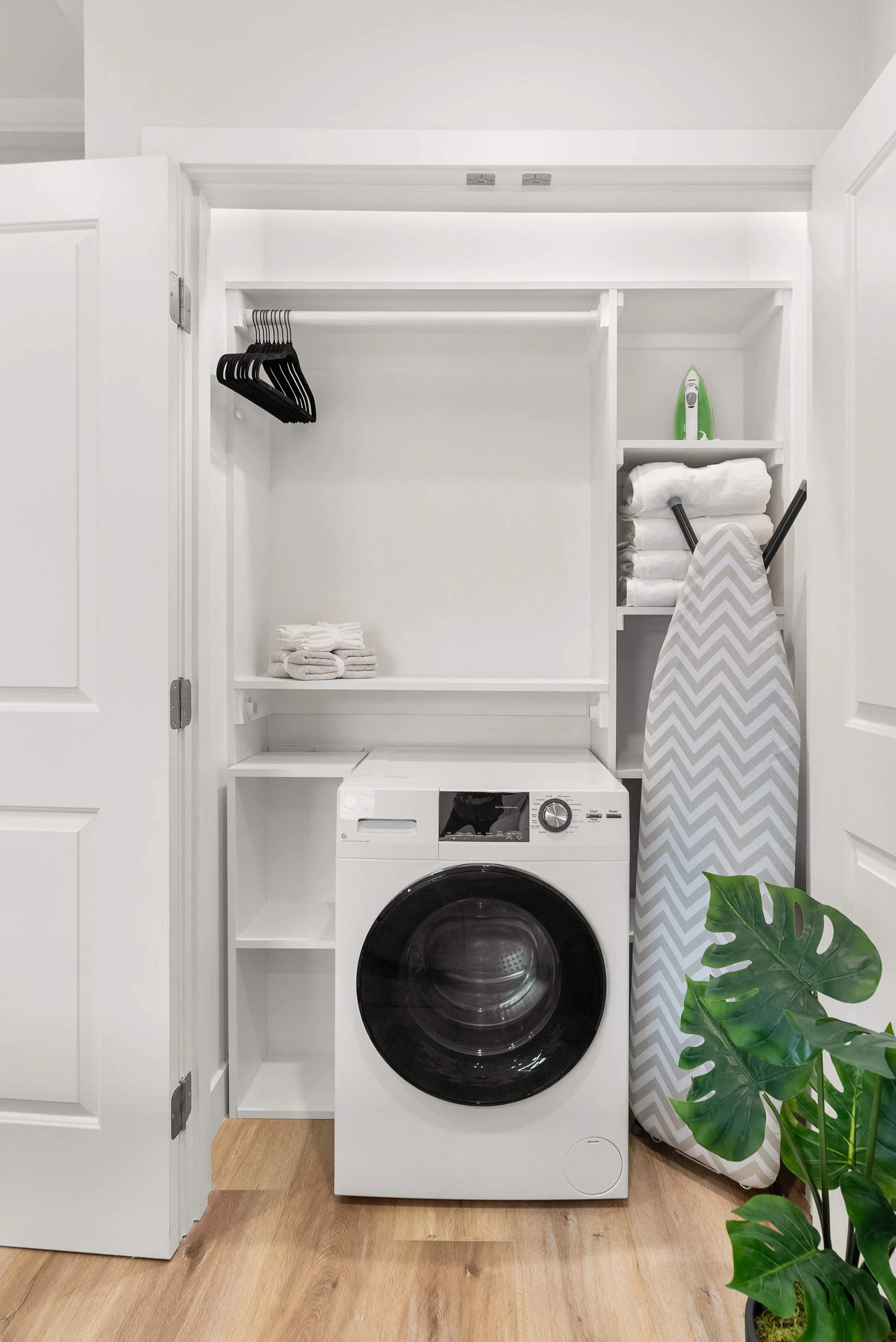 A small laundry closet with a front-loading washer, shelves with towels and an iron, an ironing board with a chevron pattern, a houseplant, and a wall-mounted iron.