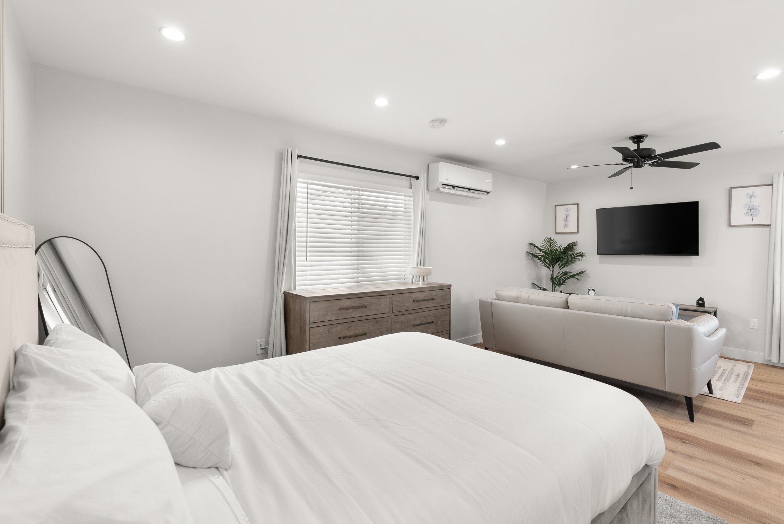 A bright, modern bedroom with white walls and ceiling, featuring a bed with white bedding on the left, a gray dresser near a window with white blinds, and a sitting area with a white sofa facing a wall-mounted TV on the right, decorated with framed a