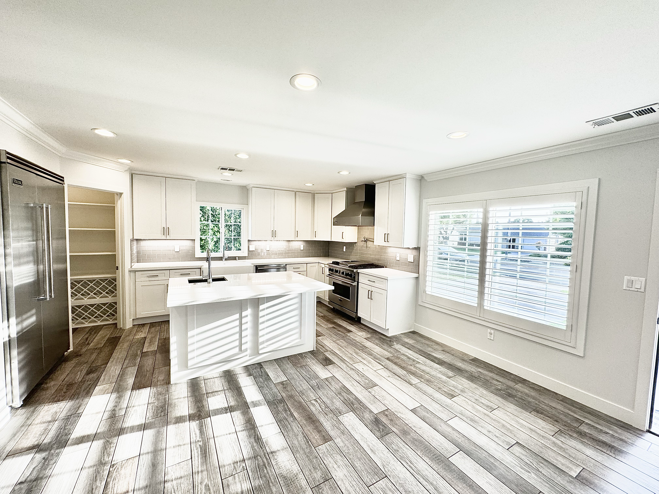 Bright, modern kitchen with white cabinets, a central island with a sink, stainless steel appliances including a refrigerator and oven, large windows with white shutters, gray backsplash, and wood-look flooring.