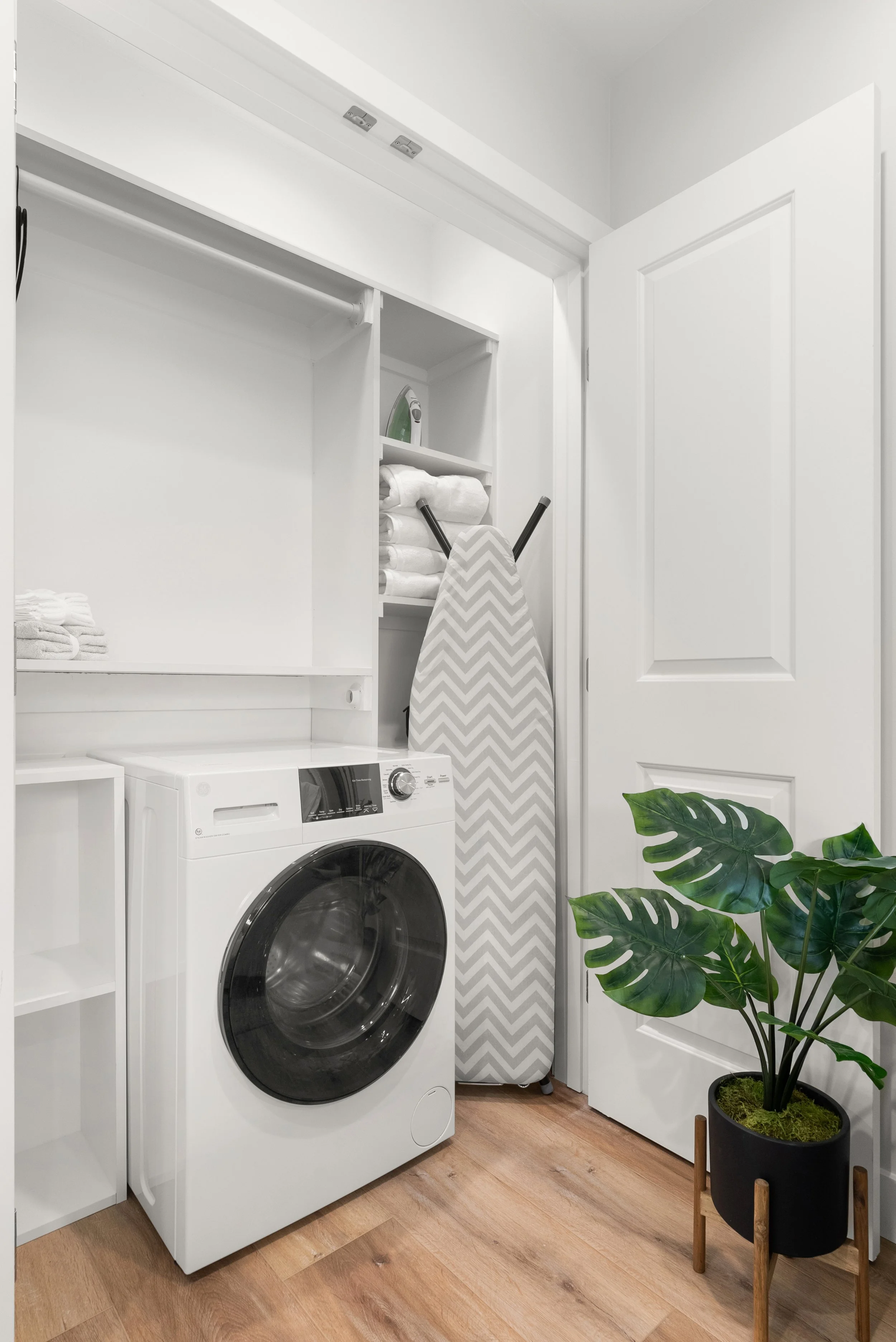A laundry room with a front-load washing machine, shelves with folded towels, an iron resting on a shelf, an ironing board with a chevron pattern, and a potted plant with large green leaves.