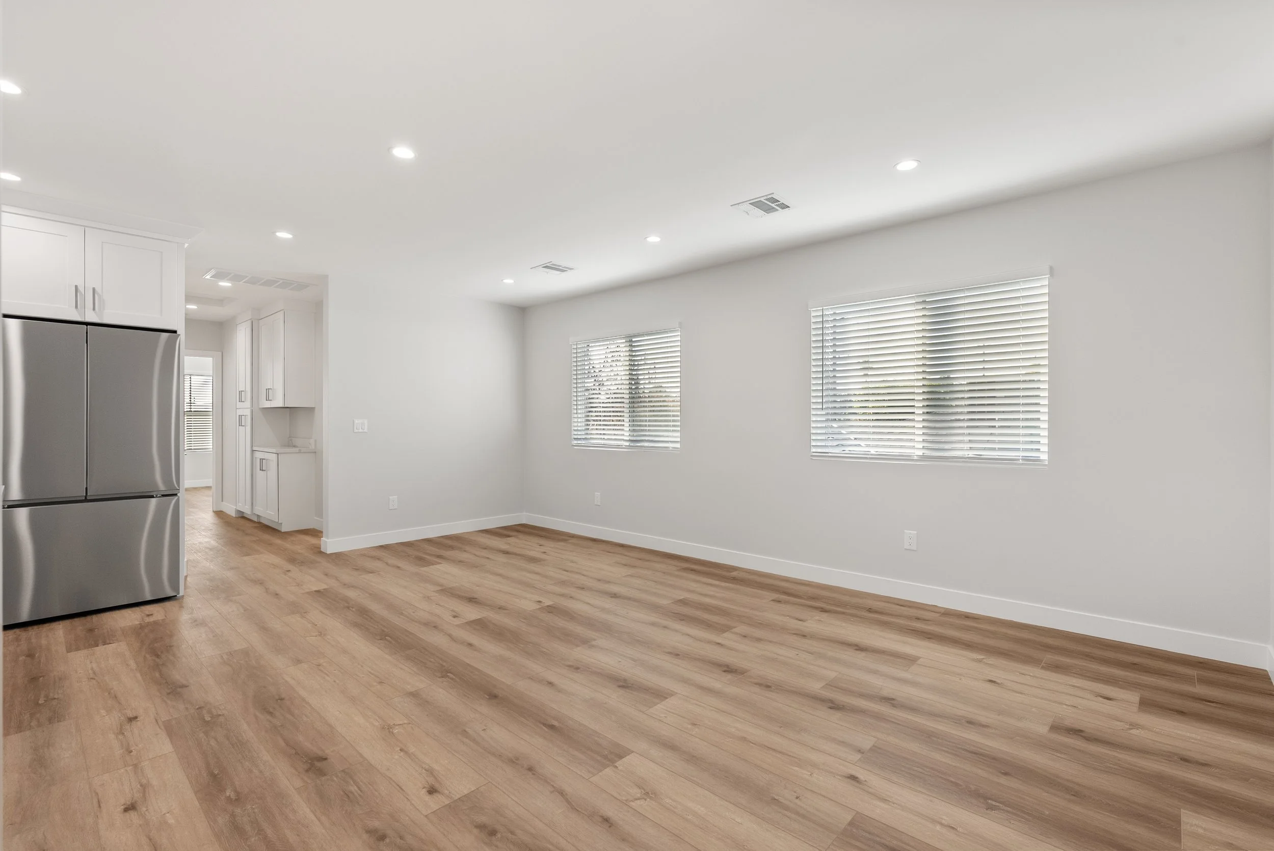 Empty living room and kitchen with white walls, hardwood floors, and windows with blinds.