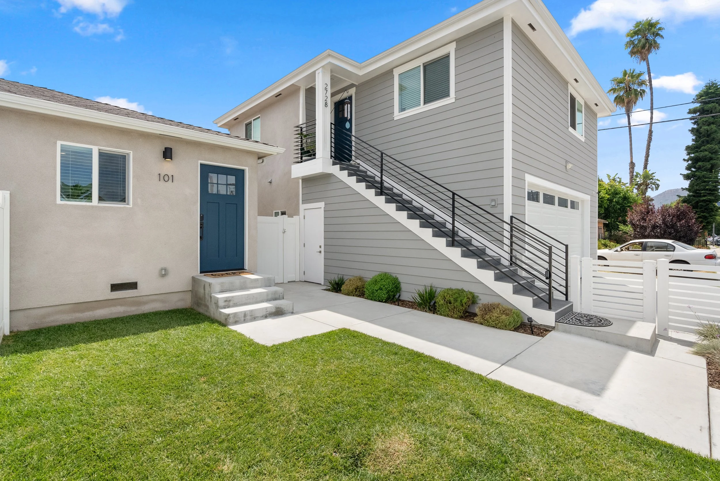 Modern two-story house with exterior stairs, gray siding, white trim, and a front yard with green lawn and plants. The house has a garage door and a blue front door with steps leading up to it.