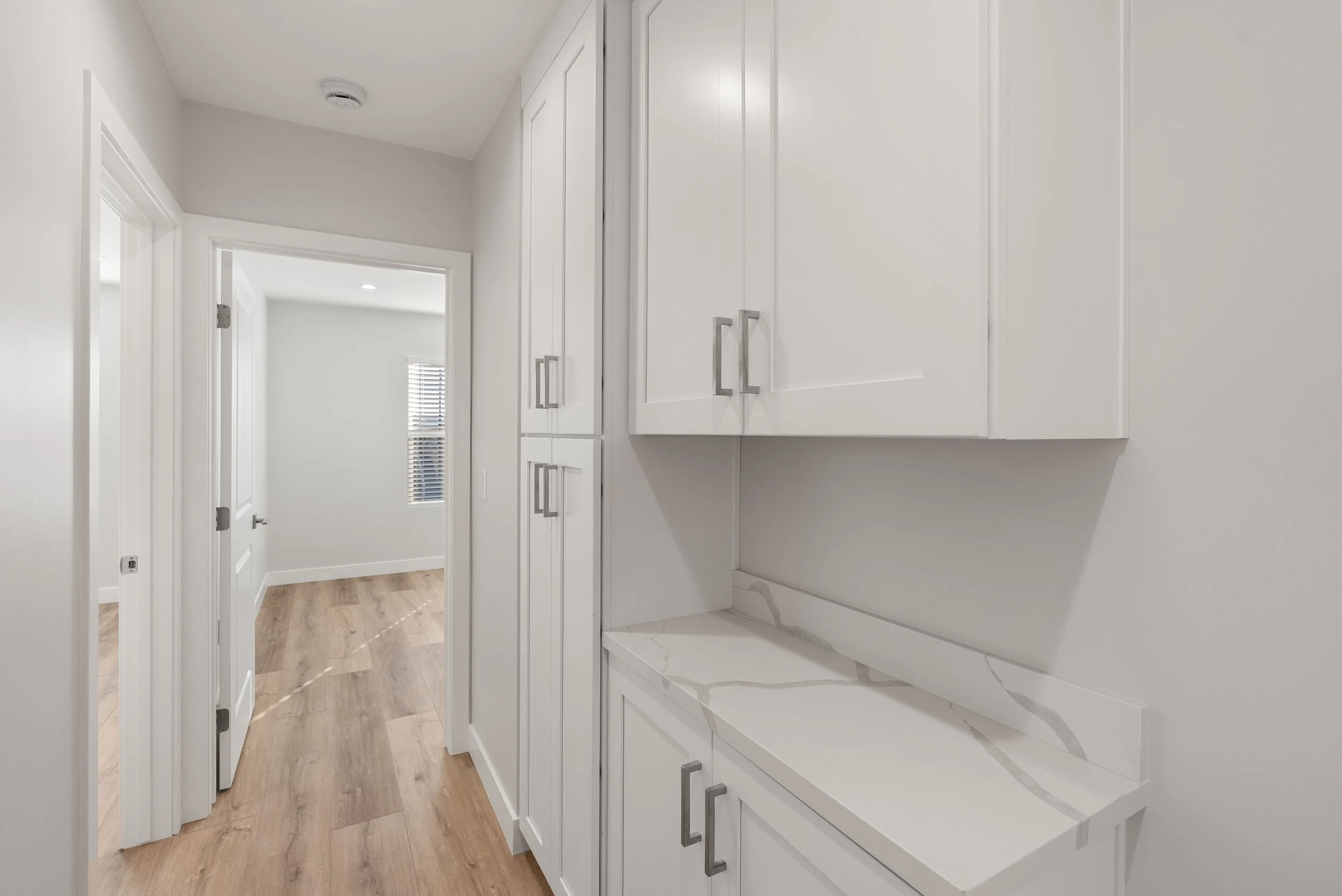 Interior view of a bright hallway with white cabinets and wood flooring, leading to a room with a window.