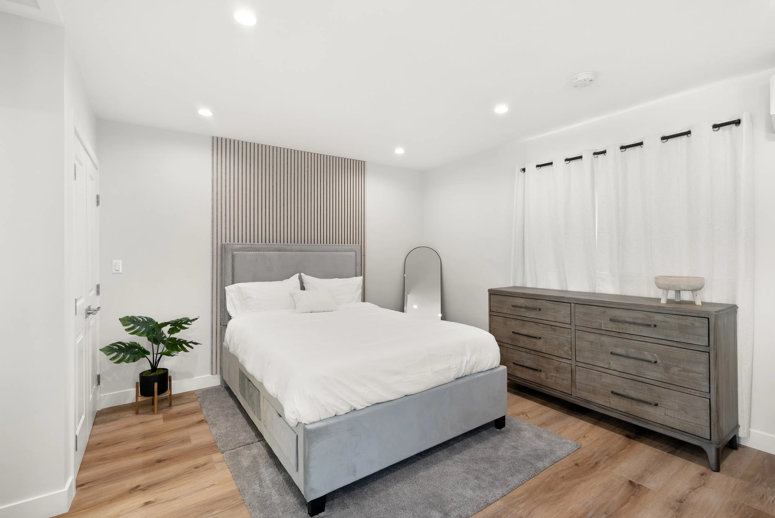 A modern bedroom with a gray upholstered bed, wooden dresser, potted plant, full-length mirror, and white curtains.
