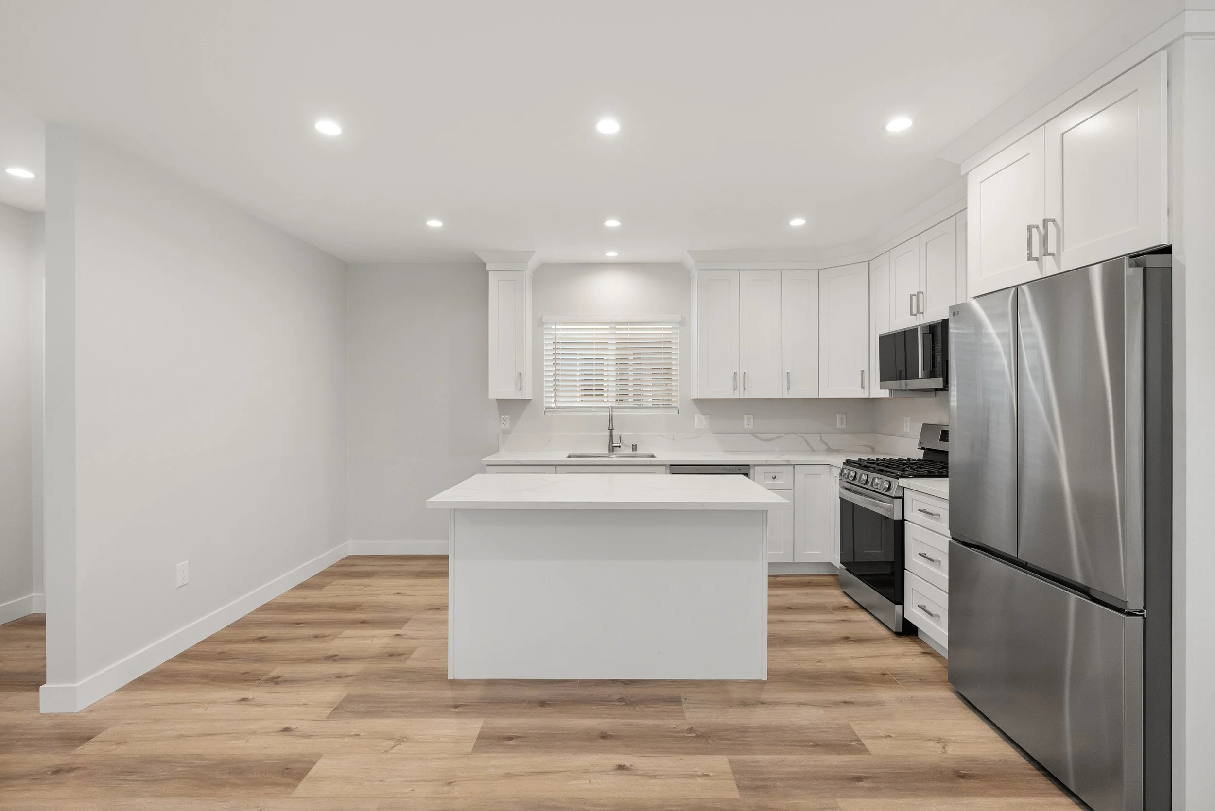 Modern kitchen with white cabinets, stainless steel refrigerator, stove, microwave, and island with white countertops, and hardwood flooring, small window with blinds.