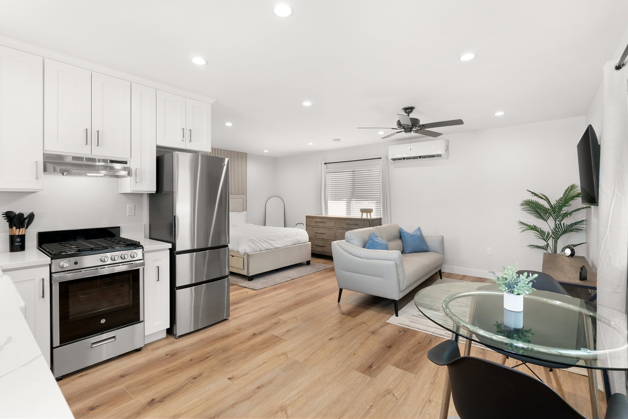 Open-concept living space with kitchen, bedroom, sitting area, and dining table, featuring white walls, wood flooring, and modern furniture.