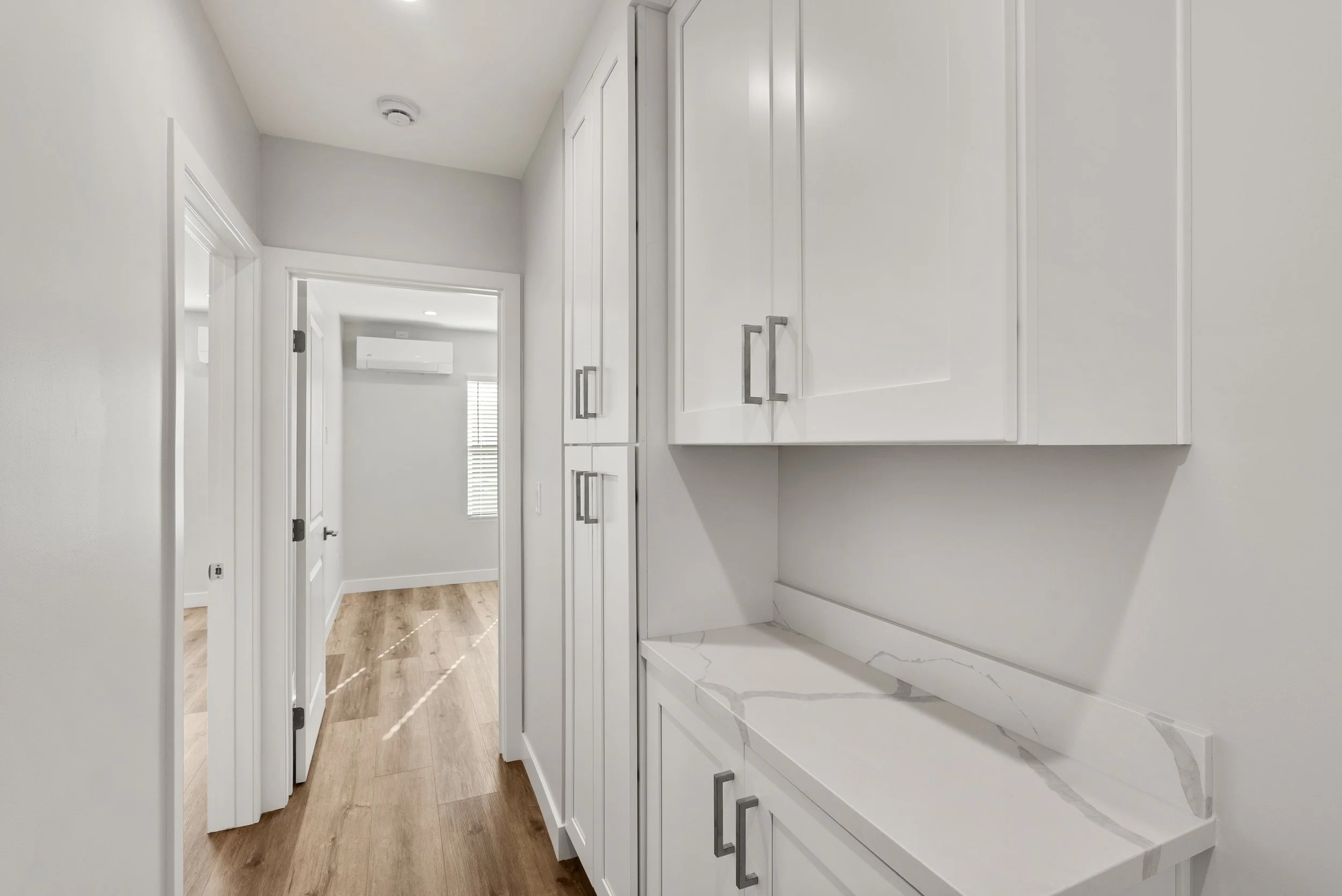 Hallway with white cabinets and a marble countertop on the right, wood flooring, and a door leading to a room with an air conditioning unit and window with blinds.
