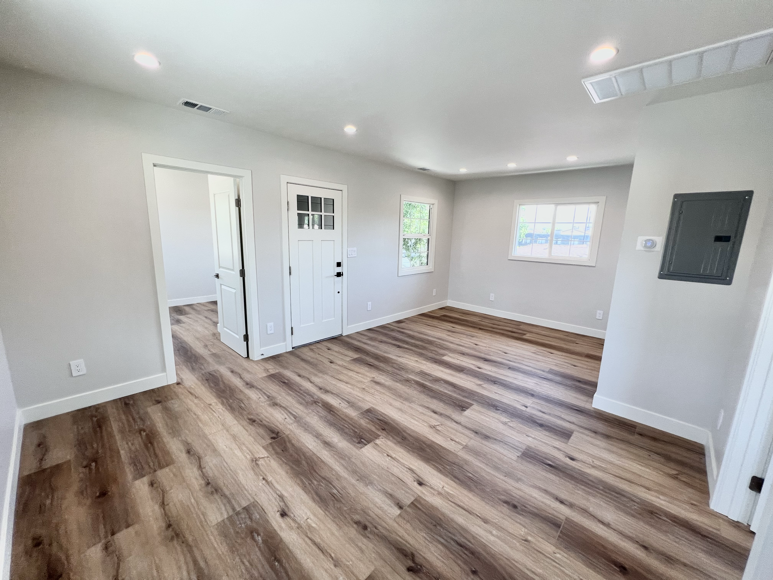 Empty living room with white walls, wood flooring, two open doors, and two windows allowing natural light.
