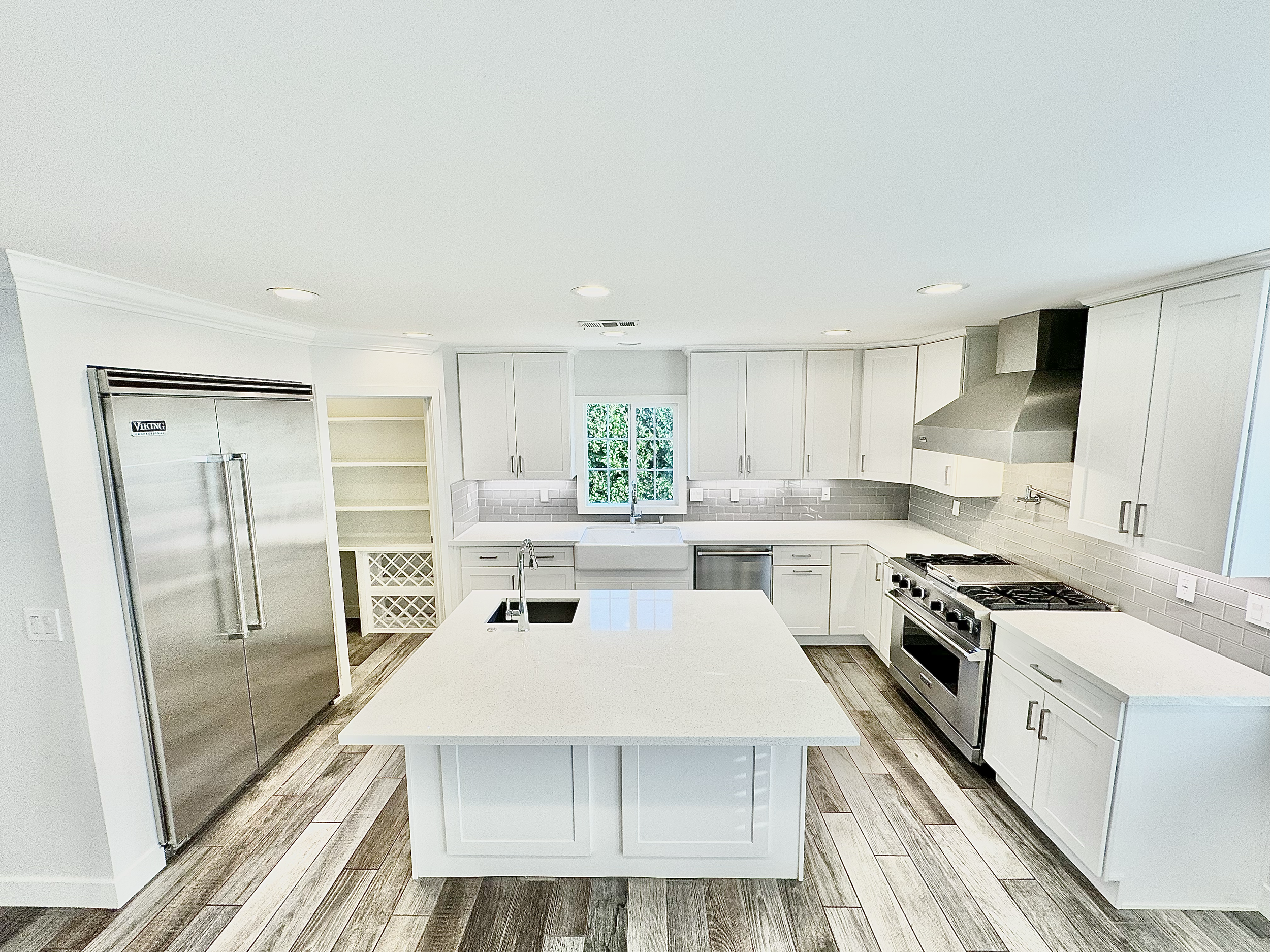 Modern white kitchen with island, stainless steel appliances including refrigerator, oven, and range hood, white cabinets, gray tile backsplash, wood flooring, window above sink, and empty open shelves.