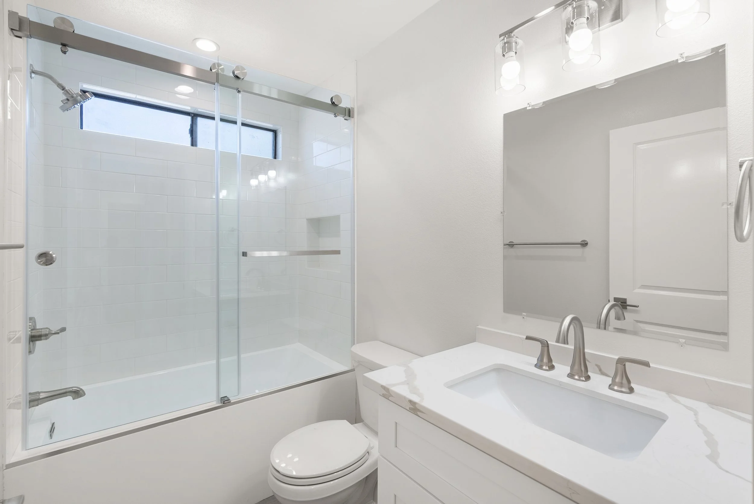 Bathroom with a glass shower enclosure, white tiled walls, a white vanity with a marble countertop, a rectangular mirror, and modern fixtures.
