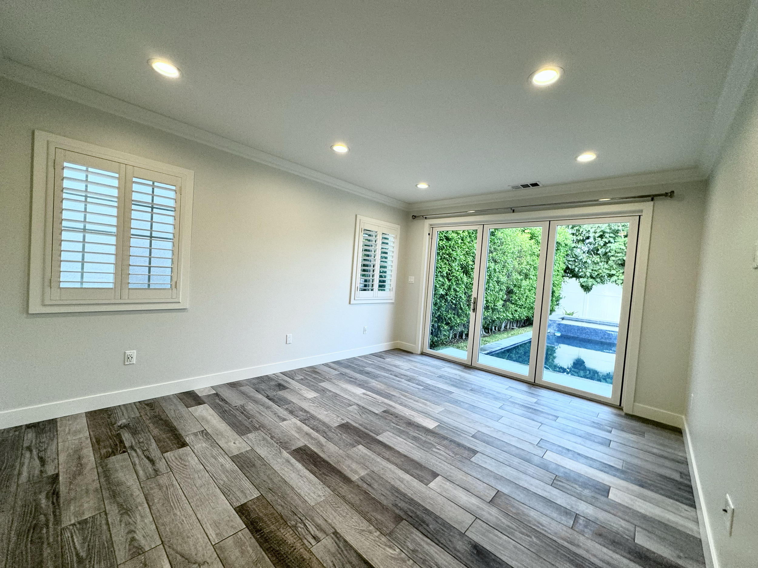 Empty living room with wood-look flooring, white walls, two small windows with shutters, sliding glass doors leading to backyard with swimming pool and greenery, ceiling lights, and a curtain rod above the doors.