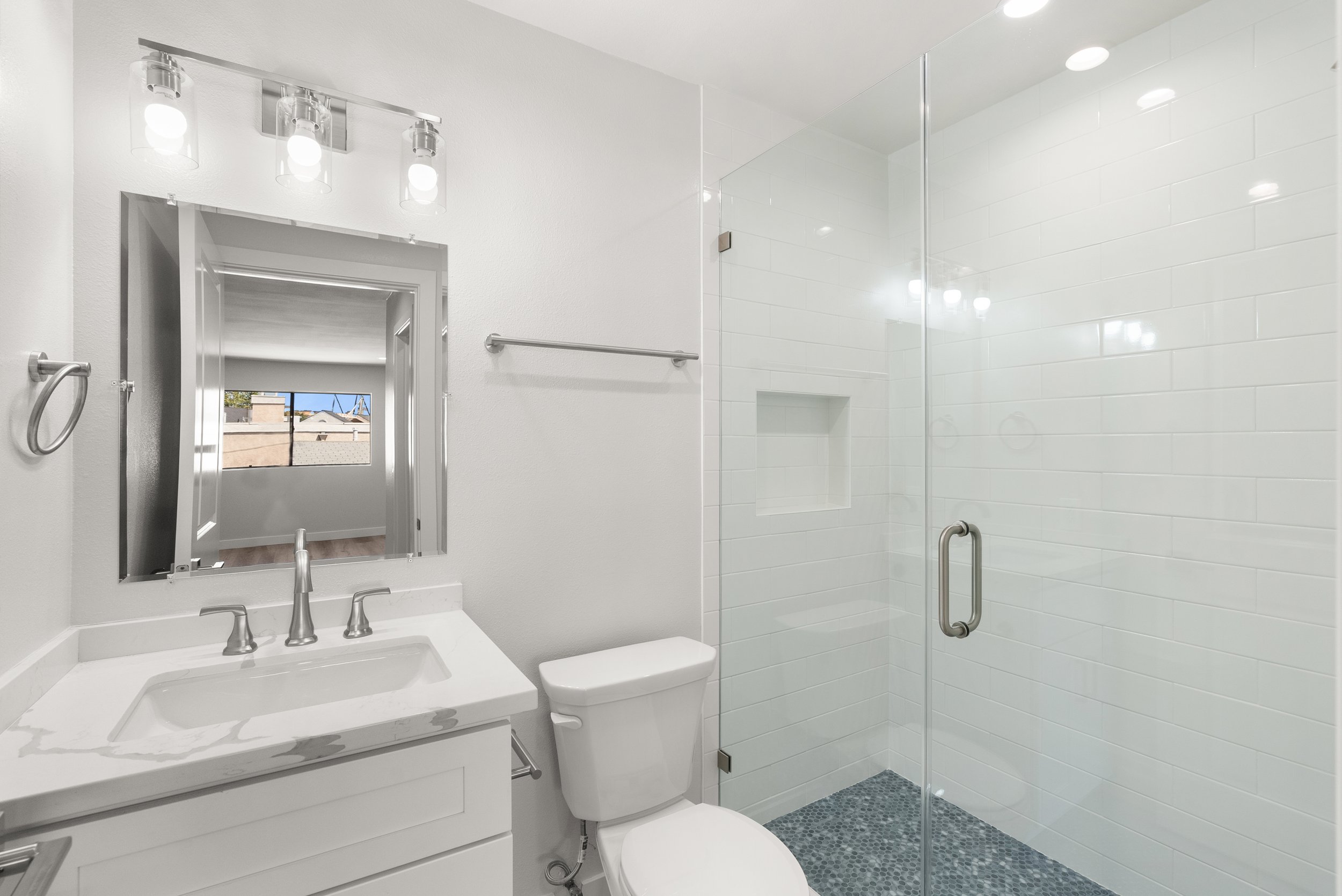 Modern white bathroom with a vanity, mirror, mirror lights, toilet, and a glass shower with white tiles and a built-in shelf.